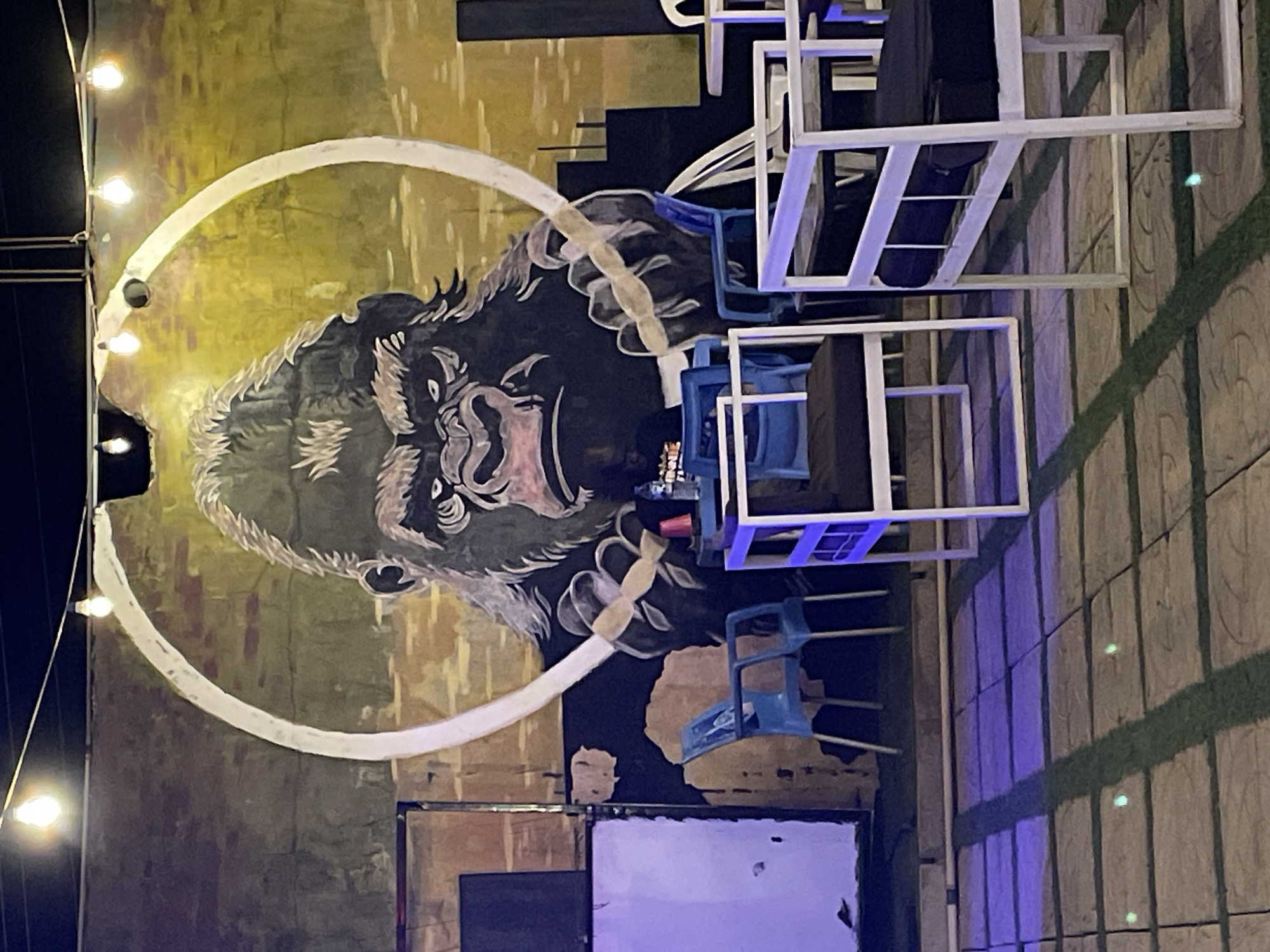 The image depicts a mural of a gorilla head with a white halo-like circle around it, located in Jos, Nigeria. The mural is painted on a textured wall with varying shades of green, brown, and black. A string of lights is visible along the left side of the mural. To the right of the mural are white framed structures and blue chairs arranged, likely part of an outdoor seating area. A section of the wall below the mural appears to be covered in dark and light painted rectangles, potentially another element of the mural or a separate design. The ground adjacent to the seating area is paved with light-colored tiles with dark green artificial turf. The lighting suggests it is nighttime.