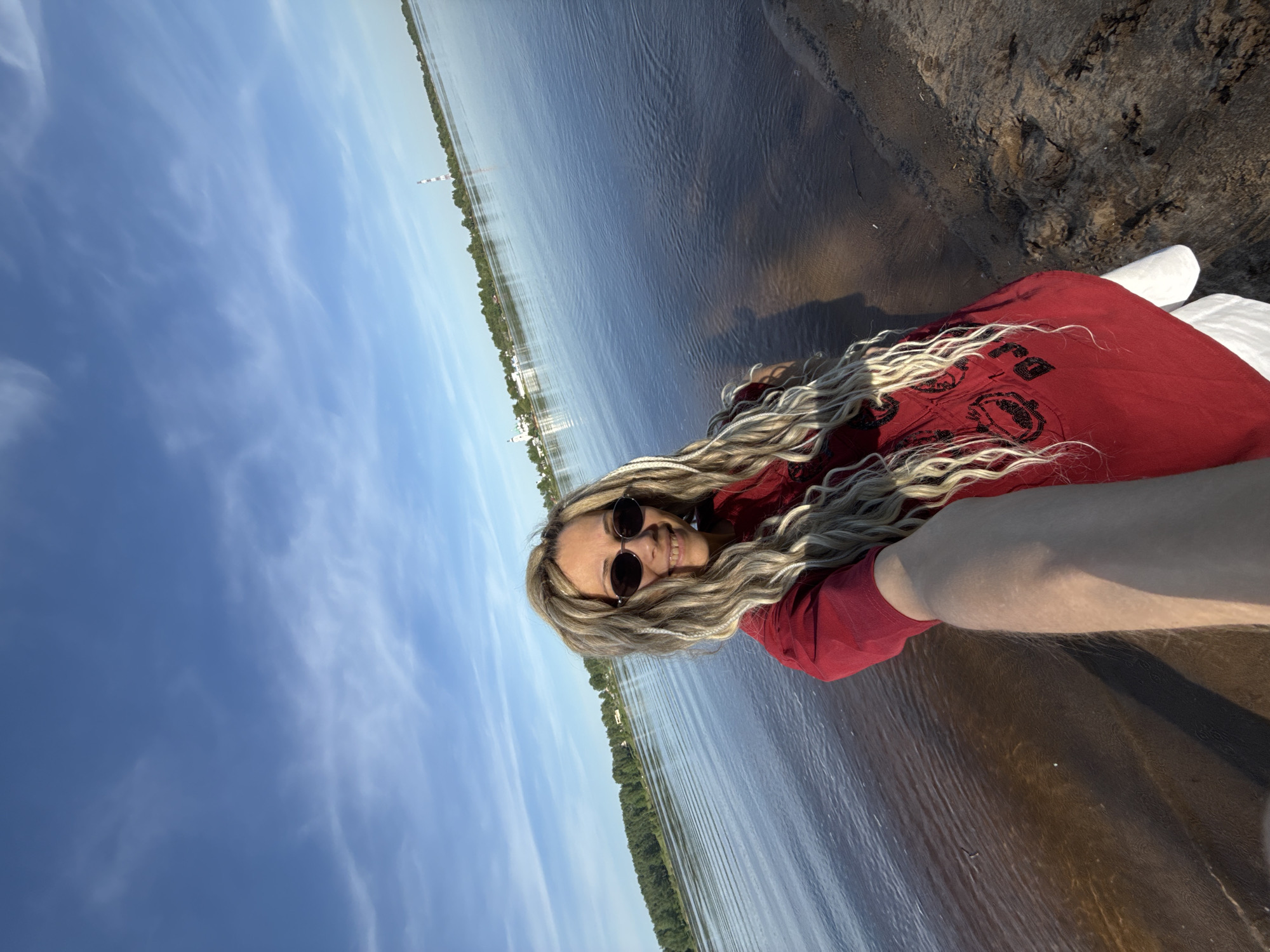 A selfie shot depicts a woman lying on a beach adjacent to water, possibly a river or lake. The location is likely near Шебунино, Russia, based on the provided context. The woman has long blonde hair, wears sunglasses, and is dressed in a red t-shirt. She smiles slightly, looking at the camera. The background includes a blue sky with some clouds, the waterline, vegetation on the opposite bank, and a sandy/soil beach area. The perspective of the photo is tilted, with the woman's head closer to the top-right corner of the frame.