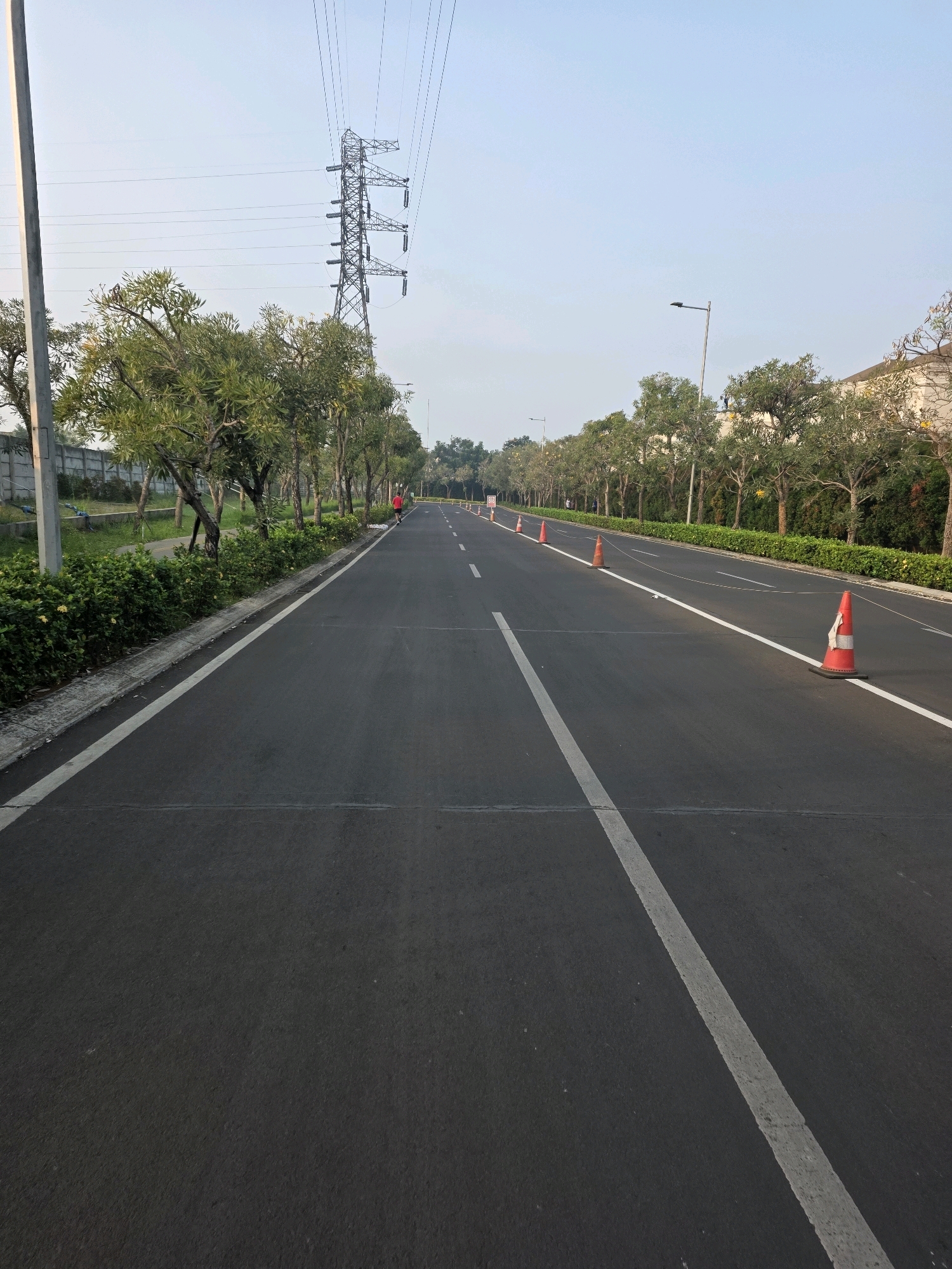 The image shows an empty asphalt road in Bojongsoang, Indonesia. The road is lined with trees and a low hedge on the right side. There are several orange traffic cones placed along the road, suggesting some form of road work or a temporary traffic alteration. In the distance, a person wearing red is jogging on the right side of the road. A tall electricity pylon is visible in the background, and the sky is clear and pale blue. The overall scene suggests a quiet, early morning setting.