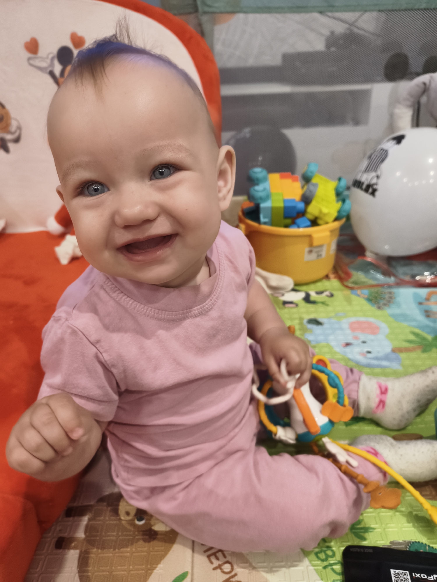 A close-up, eye-level shot shows a smiling baby with blue eyes and dark hair with a blue tint, wearing a pink t-shirt and pink pants, sitting on a play mat. The baby is in the foreground, with toys and a playpen visible in the background. The baby is holding a toy in their right hand and reaching out with their left hand. The play mat has cartoon animal designs. The background includes a yellow bucket filled with colorful building blocks, a white inflatable ball, and parts of a playpen. The scene appears to be indoors, possibly in a nursery or play area. The image composition focuses on the baby's face and upper body.