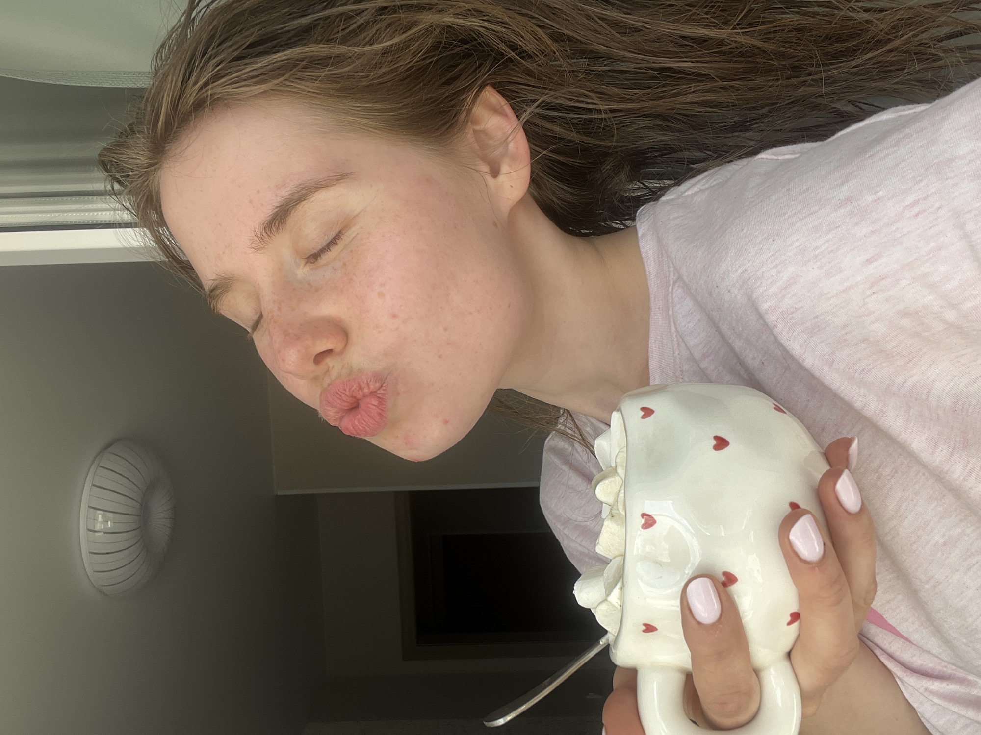 The media file shows a close-up, low-angle, portrait-oriented shot of a young woman with her eyes closed and her lips pursed in a kissing gesture. Her head is tilted back, and her hair is falling around her. She is wearing a pale pink t-shirt. In her right hand, she holds a white mug decorated with small red hearts. The mug appears to be filled with whipped cream or a similar topping, and a spoon is visible poking out of it. Her fingernails are painted a light pink.
The background is slightly out of focus, but suggests an indoor setting. To the left, a white, circular vent or fixture is visible on a light-colored wall. The lighting appears to be natural, suggesting it might be daytime. The woman's facial expression conveys playfulness or affection. The overall mood is lighthearted and intimate. No text is visible in the image. The location context is Chaikovsky, Russia.