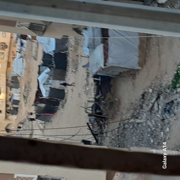 A high-angle view displays a makeshift camp or settlement. Numerous tents, some covered with white fabric, line a sandy path. Buildings are visible on the left side. Debris and rubble pile up on the right. Power lines run across the foreground, suggesting infrastructure challenges. The scene implies displacement, poverty, or emergency shelter in an arid or impoverished environment.