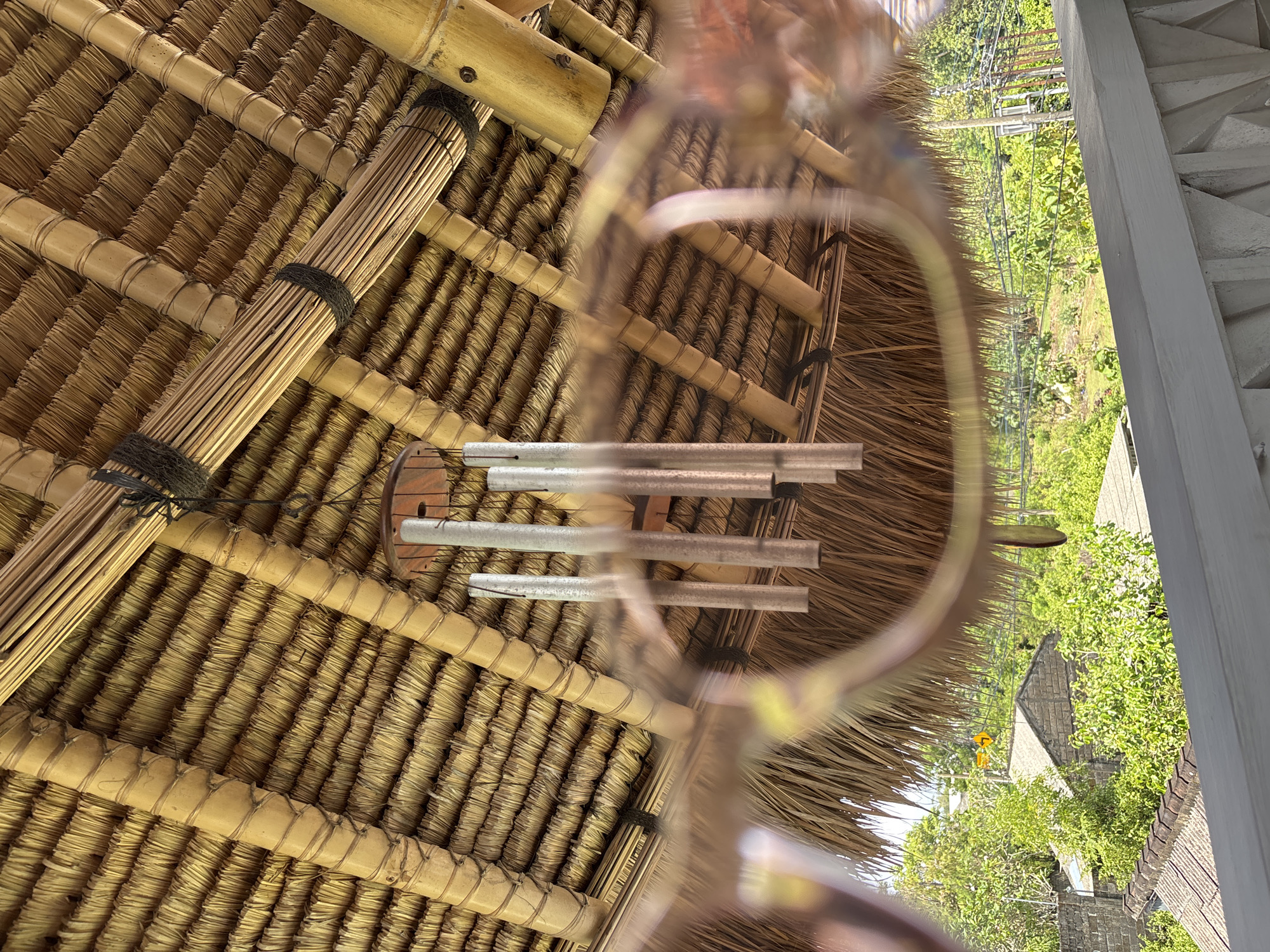 The image presents an upward view of a finely crafted thatched roof and its bamboo structural elements, set against a backdrop of a lush tropical environment. The primary perspective is rotated ninety degrees clockwise, with the outdoor horizon appearing vertically aligned.
In the foreground and midground, the intricately woven underside of a light brown thatched roof is prominent. It features numerous parallel rows of tightly bound straw or dried grass, supported by a network of horizontal and diagonal bamboo poles. These poles are secured with dark bindings. A thick bundle of smaller bamboo sticks, also fastened with dark cord, runs diagonally across the upper left section of the roof.
Suspended from the roof structure, slightly off-center to the right, is a metal wind chime, comprising several silver-colored tubes of differing lengths. Blurred, out-of-focus elements in the immediate foreground, possibly parts of another wind chime component or reflections, obscure a small portion of the scene.
Beyond the edge of the thatched roof on the right, a vibrant expanse of green foliage, including trees and bushes, fills the background. Further in the distance, parts of other buildings with grey tiled roofs and what appear to be overhead lines are visible under a bright sky. The scene is brightly lit, indicative of clear, sunny daytime weather. The materials used in the roof construction (bamboo and thatch) and the abundant tropical vegetation strongly suggest a setting in a warm climate, consistent with the provided location context of Kuta Selatan, Indonesia. No people or visible text are present in the image.