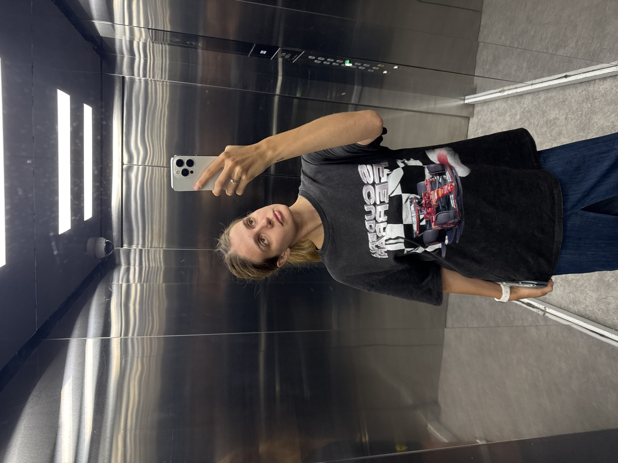 A fair-skinned female is taking a selfie with an iPhone in what appears to be an elevator. She is wearing a dark-colored graphic tee, jeans, and a white watch. The elevator's interior has a metallic finish with vertical lines and a dark control panel. A security camera is visible in the upper left corner of the frame. The location is indeterminate.