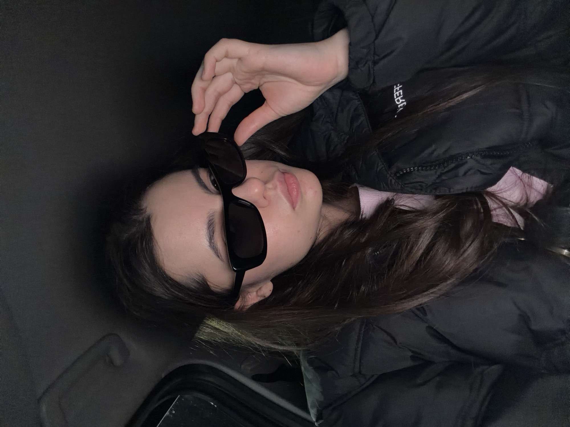 A young woman is captured in a close-up, chest-up shot, appearing to lie on her side in a dark interior space, likely a car. She is wearing a black puffer jacket, under which a light pink or purple garment is visible. Her long, dark brown hair is spread out behind her. With her right hand, she holds a pair of black rectangular sunglasses over her eyes, with only the lower part of her face, including her lips, visible. Her lips are slightly parted, adorned with light pink lipstick.
The background is predominantly dark and out of focus, with hints of a car's interior, such as a subtle grab handle on the left side of the frame. The lighting is low and appears artificial, highlighting her features against the dim surroundings. On the front of her black jacket, white text, seemingly mirrored or rotated due to the image's orientation, spells out "ICEBERG". The woman maintains a calm and composed expression while posing for the photograph. The scene is set in Chaikovsky, Russia.