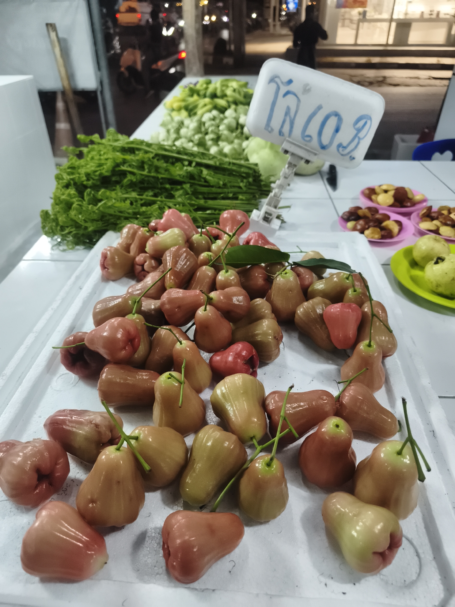 The image depicts a bustling night market stall in Baan Mae Nam, Thailand, showcasing a variety of fresh produce. In the foreground, a large white foam tray is filled with numerous rose apples (chompoo) in various stages of ripeness, ranging from deep reddish-pink to lighter green-yellow, many retaining their green stems and small leaves.
Behind the rose apples, several bundles of fresh green leafy vegetables, resembling fern shoots, are neatly arranged on a white table. Further back, piles of small, light green, roundish vegetables are visible. To the right, small pink and a green plastic plates hold different types of nuts or seeds, some dark brown and others lighter in shade.
A white sign with blue Thai writing, "โล ๔๐" (Lo 40), indicating "kilo 40" (likely 40 Baht per kilogram), stands amidst the produce. The background is blurred, showing a nighttime street scene with faint lights from cars and buildings, and a distant silhouette of a person, confirming the late hour and an active market environment. The scene highlights the availability of fresh local fruits and vegetables at an evening market.