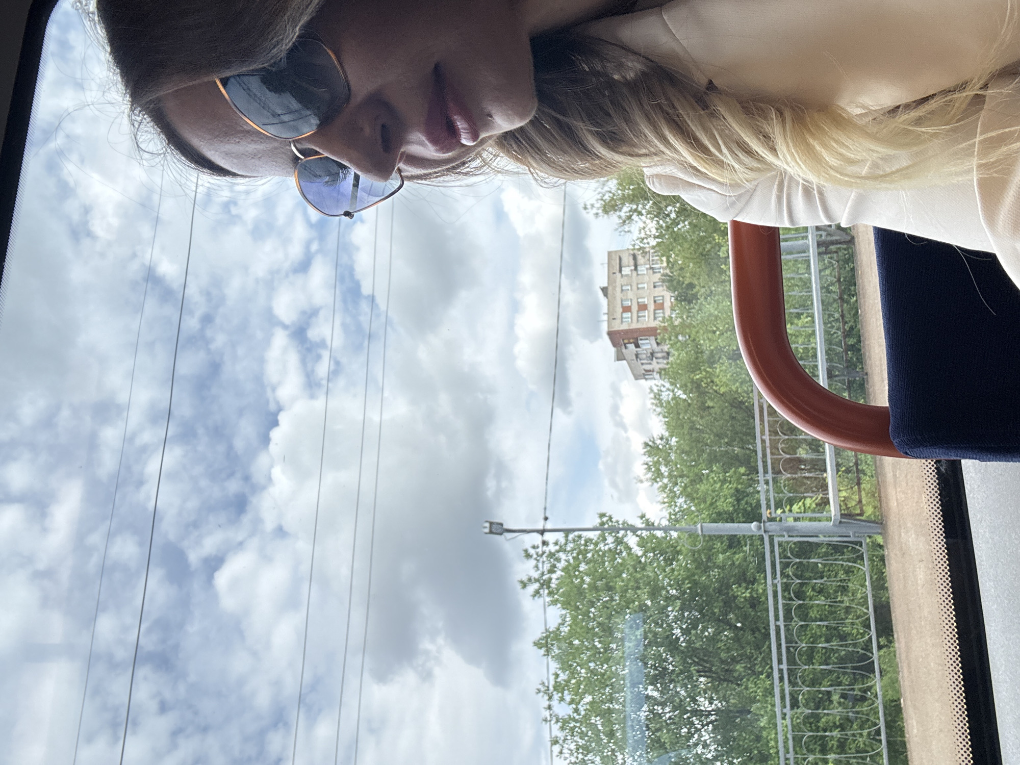 A woman with blonde hair, wearing sunglasses and a light-colored jacket, is seen in profile from the chest up, looking out of a window. The window appears to be on public transportation, possibly a tram or bus, as indicated by the presence of a red handle and a dark blue seat in the foreground. Outside the window, the scene depicts a cloudy sky with visible power lines crisscrossing the frame. Below the sky are lush green trees, a metal decorative fence, and in the distance, a multi-story building with a light-colored facade and several windows. A streetlamp is also visible amongst the trees. The overall impression is that of a journey through an urban or suburban area on an overcast day. The specific location is not discernible from the visual cues alone, but the presence of apartment buildings and greenery is typical of many cities.