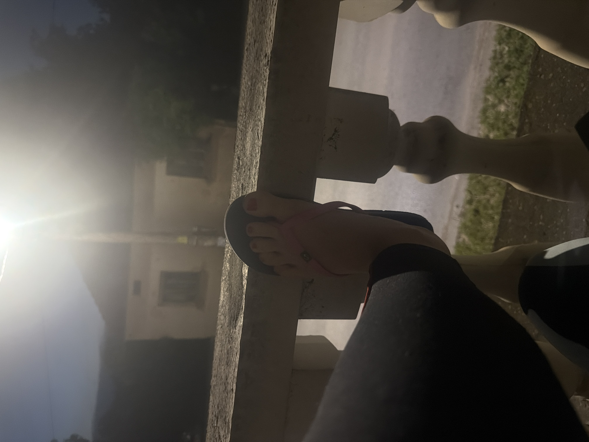 The image presents an outdoor scene captured during twilight, likely sunrise or sunset, viewed from what appears to be a balcony or porch in Iskar, Bulgaria. In the immediate foreground, a person's left foot is visible, resting on a light-colored, possibly concrete, balustrade. The foot wears a black flip-flop with a thin pink or red strap, and the toenails are painted a matching red. The person's lower leg, covered in dark leggings or pants, is also partially visible.
The balustrade runs horizontally across the midground. Beyond it, a street or paved area with patches of grass can be seen. In the background, a building with windows and walls is partially obscured by a prominent, bright light flare on the left side of the frame, which significantly overexposes that area and casts a hazy glow over the scene. Dark silhouettes of trees or dense vegetation are also discernible against the dim sky and the hazy light. The overall lighting is low, emphasizing the bright light source and giving the scene a soft, atmospheric quality typical of dawn or dusk. No visible text is present in the image.