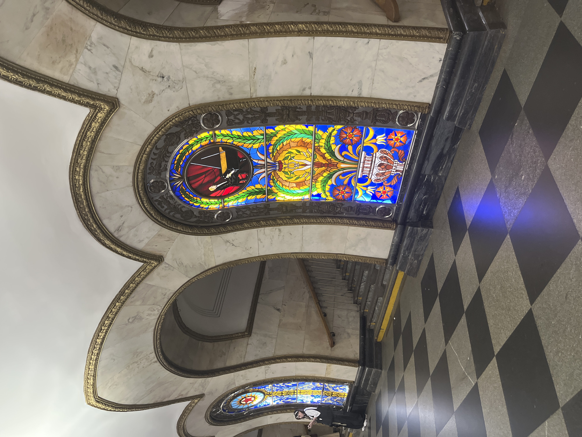 The image shows the interior of the Mayakovskaya metro station in Moscow, Russia. The station is known for its Art Deco style, featuring marble walls, ornate gold trim, and stained-glass windows. In the foreground, a black and white checkered floor leads towards a set of stairs. To the right, a large stained-glass window depicts a musician playing a piano, surrounded by floral motifs and geometric patterns. To the left, another stained-glass window, partially obscured, shows a red star within a circular design. A person wearing dark clothing and a hat stands near the left window, holding a phone. The overall impression is one of grandeur and artistic design characteristic of Soviet-era metro stations.
