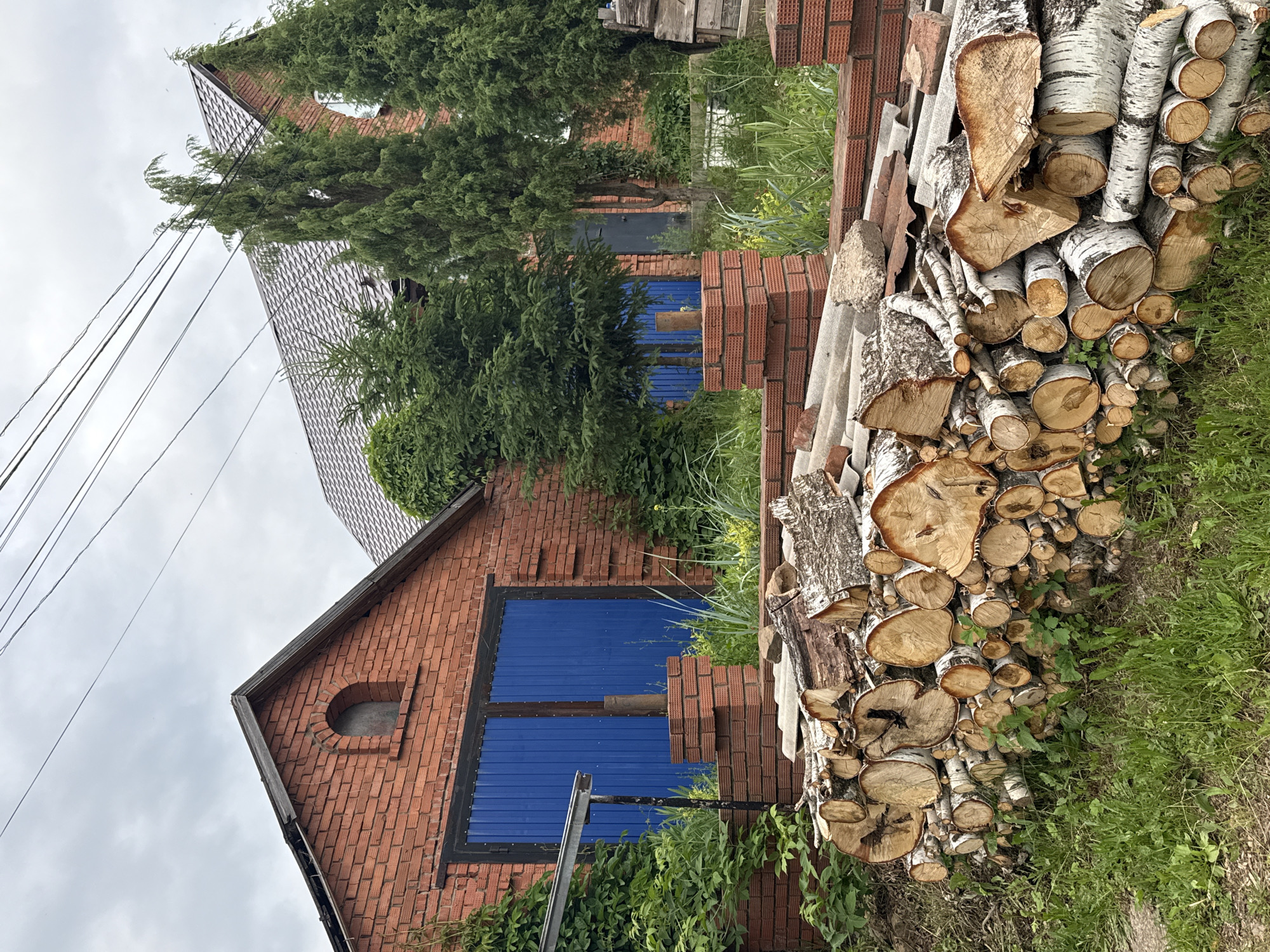 The image depicts an outdoor scene in Votkinsk, Russia, featuring a pile of chopped firewood in the foreground. The wood, primarily birch, is stacked neatly against a backdrop of red brick buildings and lush green vegetation. The larger building on the left has blue double doors, and a smaller arched window is visible above them. To the right, another brick building with a grey tiled roof is partially visible, with several tall evergreen trees in front of it. Overhead, power lines crisscross the cloudy sky. The overall impression is that of a rustic, possibly rural or suburban, setting.