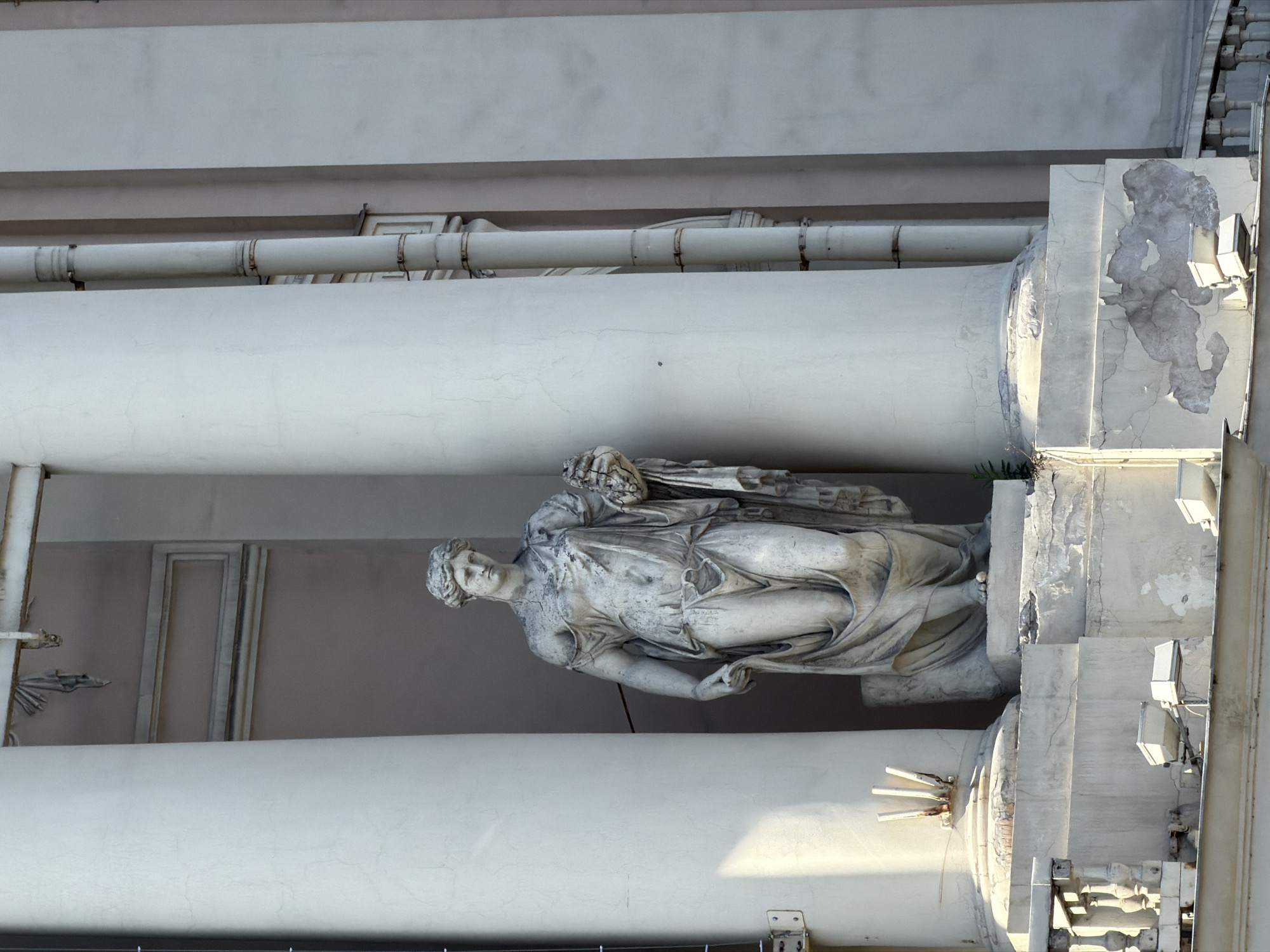 A full-length, classical-style stone statue of a female figure stands prominently between two large, vertical architectural columns. The statue, depicted in a flowing draped gown that reveals intricate folds, has her head turned slightly to her left. She holds an indistinct object, possibly a sheaf or bundle, with her right arm, resting on her shoulder. The sculpture displays visible signs of weathering, including cracks and surface discoloration.
The massive, light-colored columns flanking the statue are integral to a grand building facade, also showing age through dirt, cracks, and minor damage. Above the statue and columns, a horizontal section of the building is visible. Below this, a series of white utility pipes, secured by metal bands, runs horizontally across the upper part of the structure.
To the right of the rightmost column, the building facade shows significant gray discoloration and peeling paint. Several modern, square-shaped outdoor lights are mounted on this section, and a stone balustrade or railing is visible below. To the left, behind the left column, is a simpler wall surface featuring a recessed panel or frame. The entire scene is bathed in natural daylight, with shadows cast from the left, indicating it is daytime. The architecture suggests a historical European setting, consistent with Saint Petersburg, Russia. No text is visible in the image.
