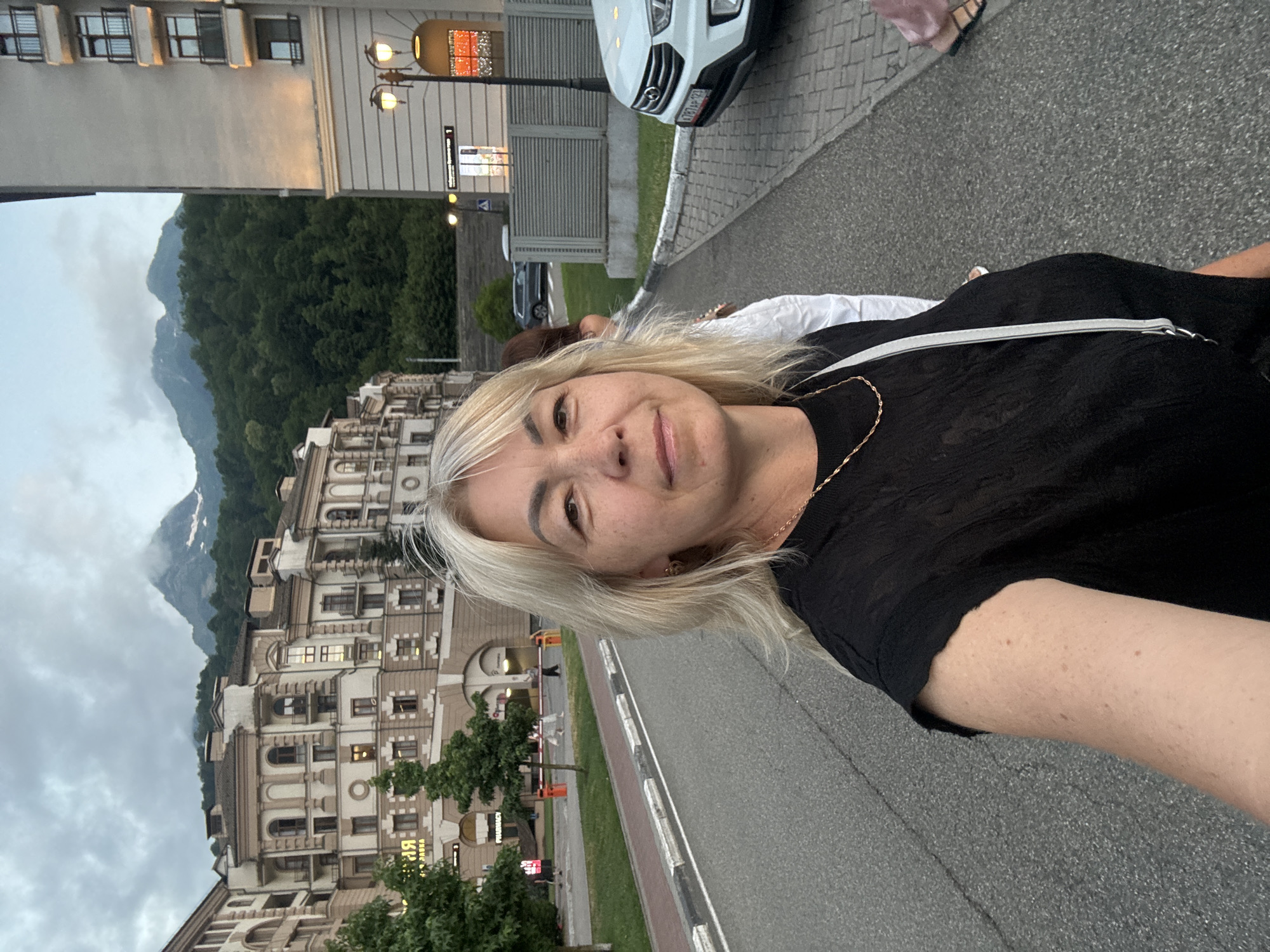 A woman with blonde hair is taking a selfie outdoors in a setting that appears to be a resort area. She is wearing a black top and a light-colored purse is slung across her shoulder. In the background, there is a large, ornate building with multiple stories and arched windows. Mountains covered in lush green trees and some snow-capped peaks rise behind the building. The sky is overcast, suggesting it might be late afternoon or evening, and the weather appears to be mild. The woman is smiling, conveying a relaxed and happy mood. The pavement in front of her suggests a public walkway or road. There is visible text on a sign on the building that reads "PHARMACY" and some Cyrillic text. The location is identified as Krasnaya Polyana, Russia, a known mountainous resort area.