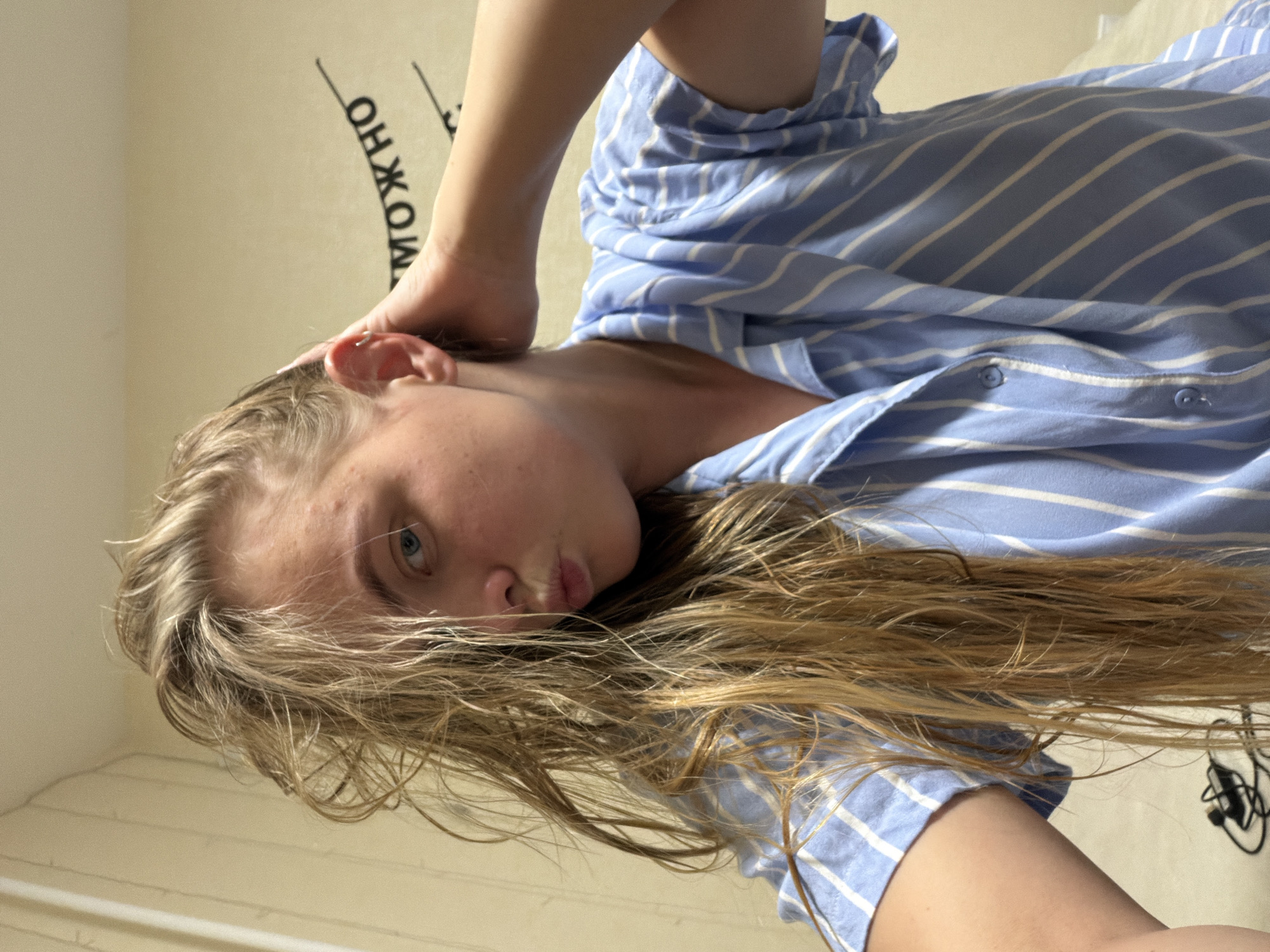 A young person with blonde hair and blue eyes, wearing a light blue and white striped shirt, is positioned close to the camera. The background includes a light-colored wall with a partially visible inscription in Cyrillic, possibly the word "Можно". Due to the limited context and lack of clear landmarks, it is not possible to determine if the location is Izhevsk, Russia, or the exact nature of the scene. The photo's angle suggests a selfie or a low-angle shot.
