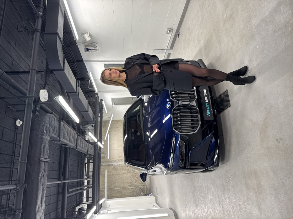 A fair-skinned woman with long blonde hair poses on a dark blue BMW X7. She wears a black blazer over a sheer top, a black miniskirt, and black tights with black suede booties. The car's front grill is visible. "Tiffany Motors" is printed on a plate to the side of the car. The setting is a showroom with black ventilation equipment in the corner and bright overhead lights. The floor and walls are gray concrete and white painted brick, respectively.