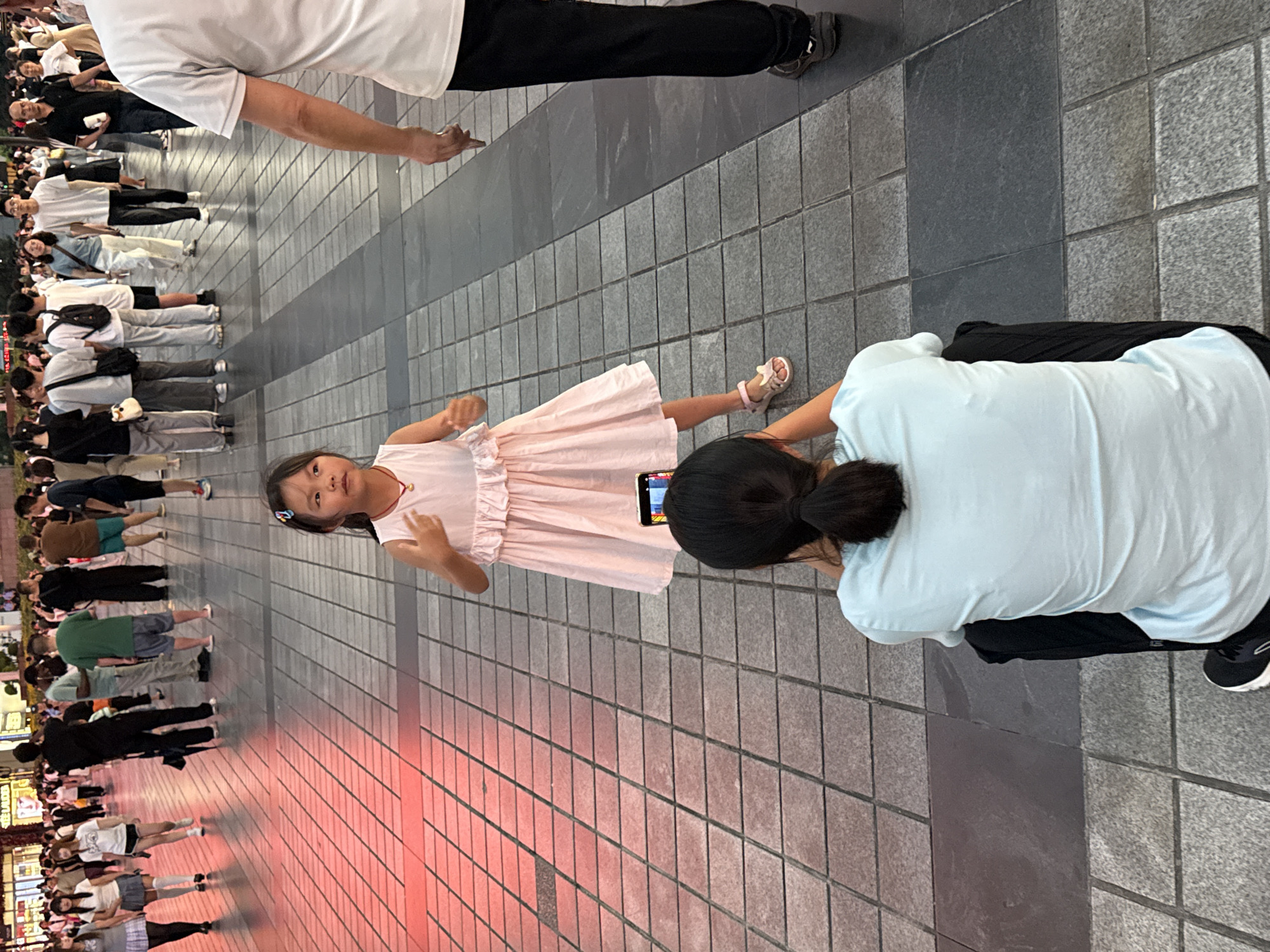 An over-the-shoulder shot captures a young girl in a pink dress posing for a photo in Yuzhong District, China. The girl stands on a paved walkway, her hands held up. A woman is crouched in the foreground, holding a smartphone to take the picture. The background shows a crowd of people walking, dressed in casual clothing. The lighting suggests it is late afternoon or early evening, with artificial lights also visible. The ground is paved with square tiles.