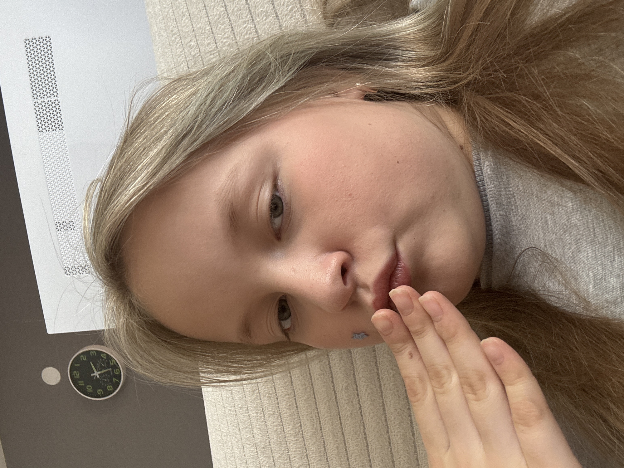 A blonde-haired person is lying down indoors. A hand is partially covering their mouth. A clock is visible on the wall, showing the time as approximately 11:20. The presence or lack of data means the image cannot be placed in the city of Izhevsk, Russia, with certainty. There is a computer panel, and a textured beige wall.