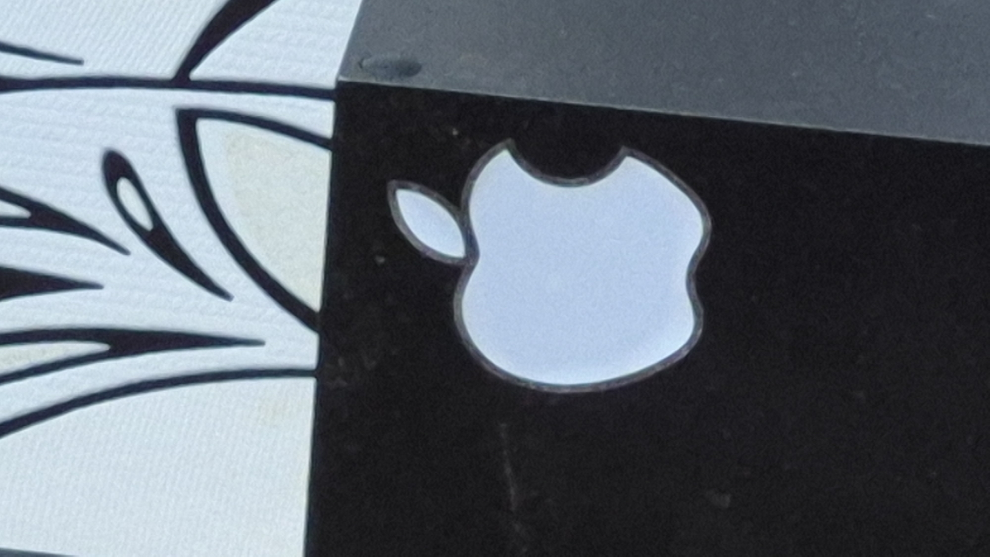 The image displays a close-up of an Apple logo on a black surface, likely a product or packaging. The logo is a light blue silhouette of an apple with a bite taken out of it, outlined in a darker shade. Above the black surface, a white background is visible with some abstract black lines. The overall image is slightly grainy. The location in Neftegorsk, Russia, is not depicted in the image.