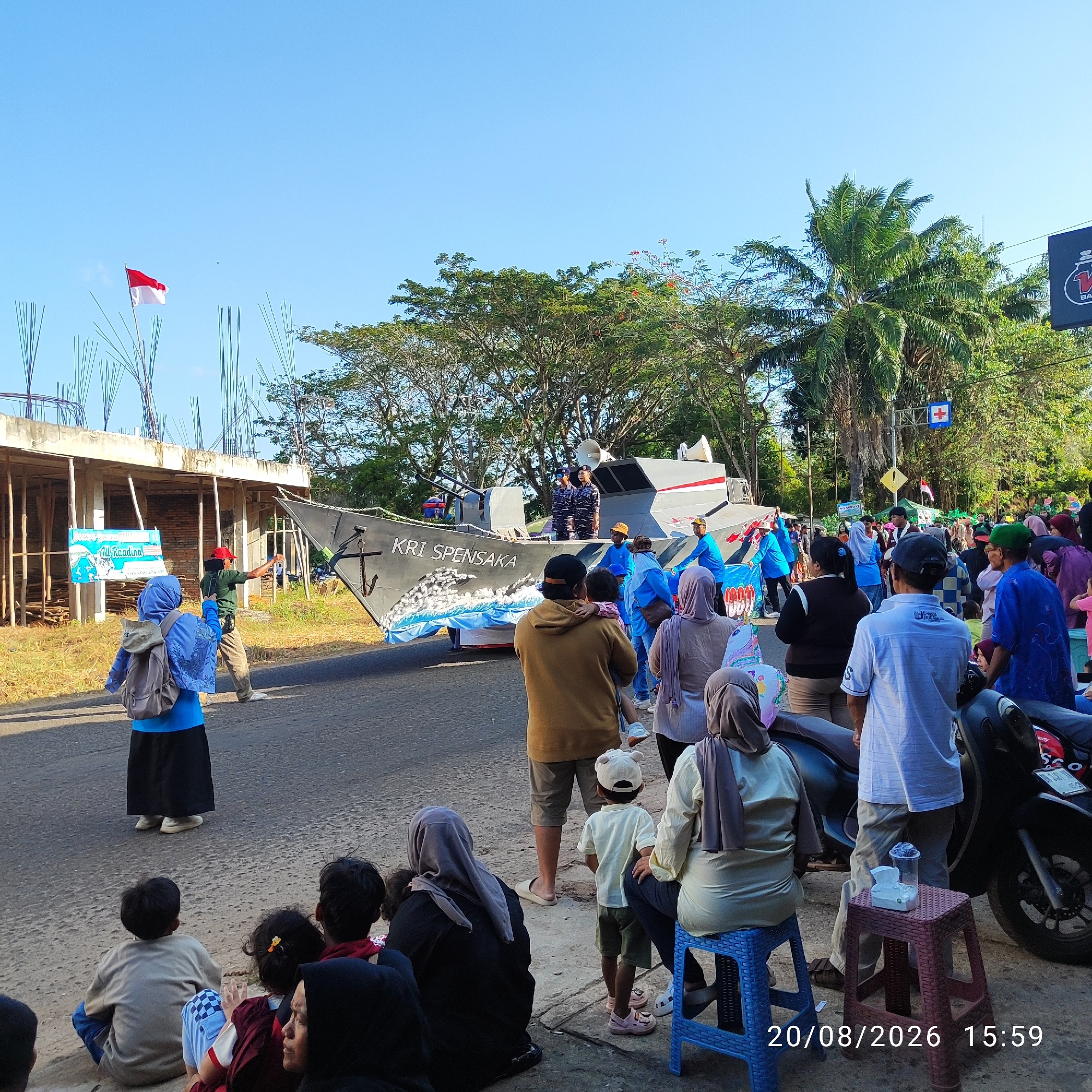 In Buding, Indonesia, a street scene depicts a parade or event with a replica of a ship named "KRI SPENSAKA" as the central float. The ship is gray with white and red accents and features a drawing of waves at its base. On the ship, two individuals in navy uniforms stand at attention. A crowd of onlookers, including children and adults, are gathered along the roadside, many in casual attire and some wearing headscarves. Some people are sitting on the curb, while others are standing. A woman in a blue headscarf and backpack is taking a photo of the float with her phone. To the left of the road, construction scaffolding and unfinished buildings are visible. Trees and palm trees line the background, and the sky is clear and blue. The timestamp "20/08/2026 15:59" is present in the lower right corner.