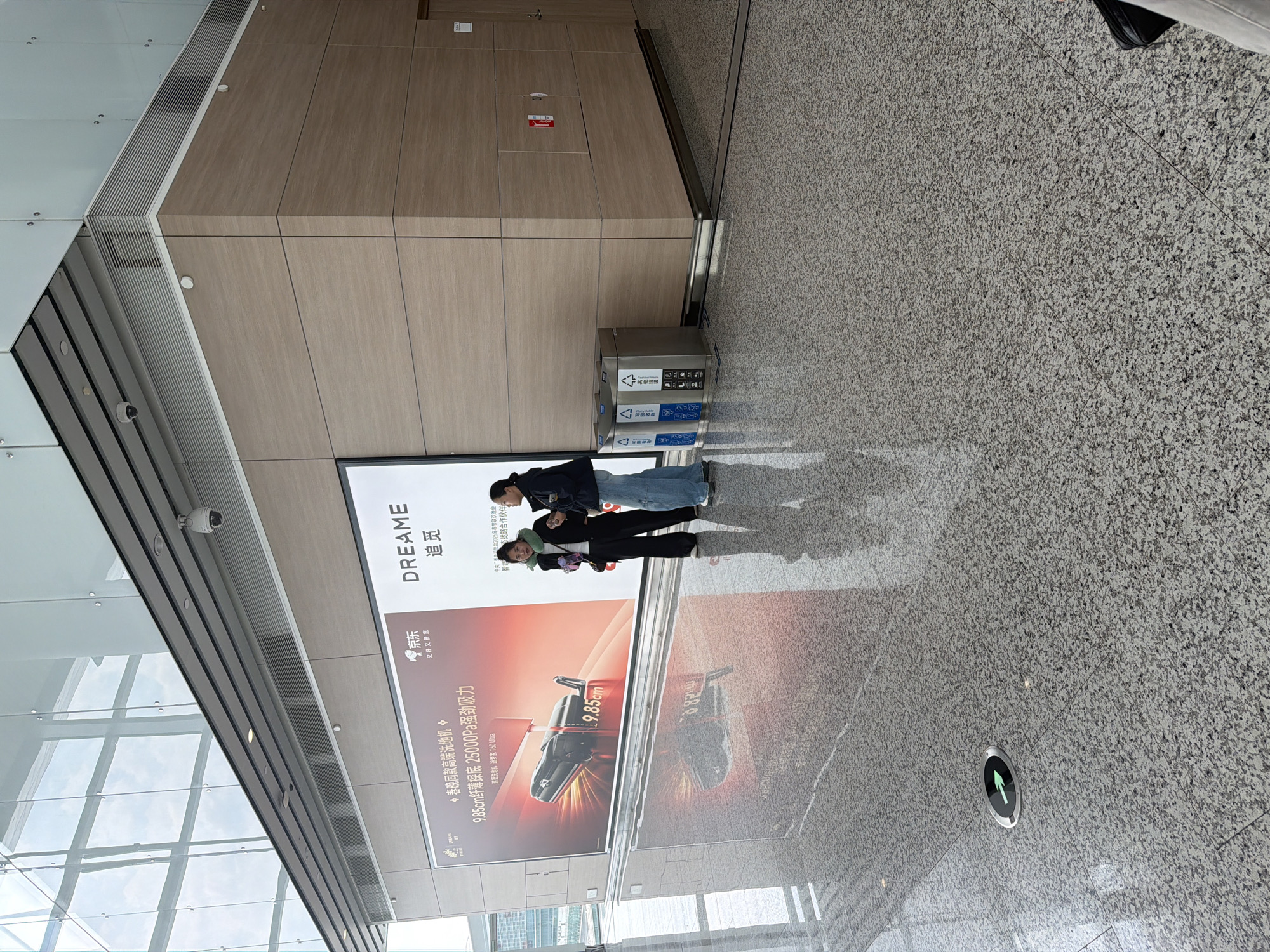 The image captures an indoor scene within a modern public space, possibly an airport or large commercial building, featuring two individuals, a large advertisement display, and trash bins. The setting is characterized by clean, contemporary architecture with light wood-paneled walls, a textured grey terrazzo-style floor with reflective qualities, and expansive glass panels reflecting the sky and interior structures. A security camera is mounted on the ceiling.
Two individuals are standing near a prominent advertisement. One person wears a dark jacket over light blue jeans, while the other wears a dark jacket, a light green hood, and dark pants. They appear to be casually standing or waiting, with their reflections visible on the polished floor.
The main object is a large digital advertisement displaying the brand "DREAME" (追觅), with the slogan "又好又便宜" (good and cheap), associated with Jingdong (京东), a Chinese e-commerce company. The ad features a product, a "高端洗地机" (high-end floor washer), described as "9.85cm纤薄探底 25000Pa强劲吸力" (9.85cm slim deep reach, 25000Pa strong suction), and references its appearance at the "春晚" (Spring Festival Gala), specifically mentioning "2006年春节联欢晚会" (2006 Spring Festival Gala) and being a "战略合作伙伴" (strategic partner).
In front of a wood-paneled wall, two stainless steel trash bins are visible, labeled with Chinese characters "可回收垃圾" (Recyclable waste) and "其他垃圾" (Other waste), along with recycling symbols. A circular floor-level light fixture with a green arrow pointing forward is embedded in the floor.
The lighting suggests it is daytime, with natural light entering through the large windows, contributing to the bright and open atmosphere. The specific location cues, primarily the Chinese text on the advertisements and bins, indicate the setting is in a Chinese-speaking region.