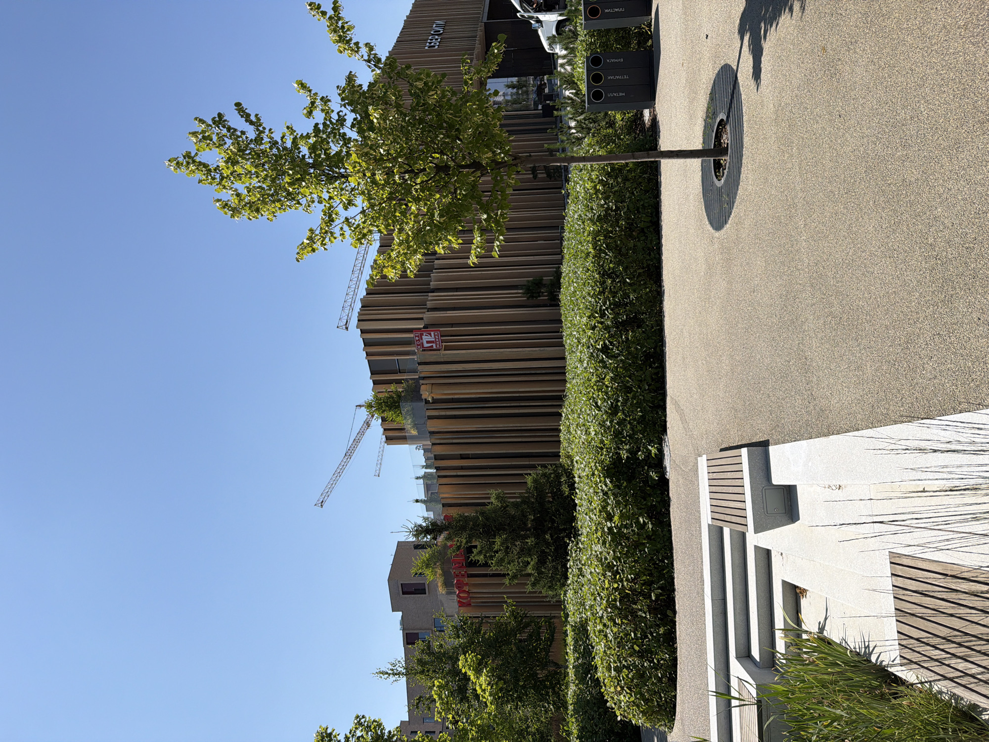 The image depicts a bright, sunny day in a modern urban park area. The foreground features a paved walkway with a speckled texture, bordered by a low, multi-tiered seating area constructed from light-colored stone and wooden slats. Greenery, including ornamental grasses and shrubs, is interspersed throughout this area.
To the left of the walkway, a dense, well-maintained hedge forms a visual boundary. Beyond the hedge, several trees with green foliage are visible.
Dominating the midground is a contemporary building with a facade clad in vertical wooden slats. A prominent red sign with white lettering, partially obscured by trees, reads "КОПТЕИ". The building also features windows and what appears to be a rooftop garden. Two construction cranes are visible in the background, suggesting ongoing development in the area. A sign on the right side of the building reads "СБЕР СИТИ".
In the right midground, a young tree stands on the paved area, with a circular protective grate around its base. Adjacent to the tree are three recycling bins, labeled in Cyrillic: "МЕТАЛЛ", "ТЕТРАПАК", "БУМАГА", and "ПЛАСТИК". A white van with its door open is parked to the right of the building.
The sky is clear and bright blue. The overall impression is of a newly developed or renovated public space designed for recreation and possibly commerce. The presence of the "СБЕР СИТИ" sign suggests this location is likely within a Sberbank-related complex or development. Given the Cyrillic signage and the architectural style, the location is likely in Moscow, Russia, possibly in a business or mixed-use district undergoing construction.