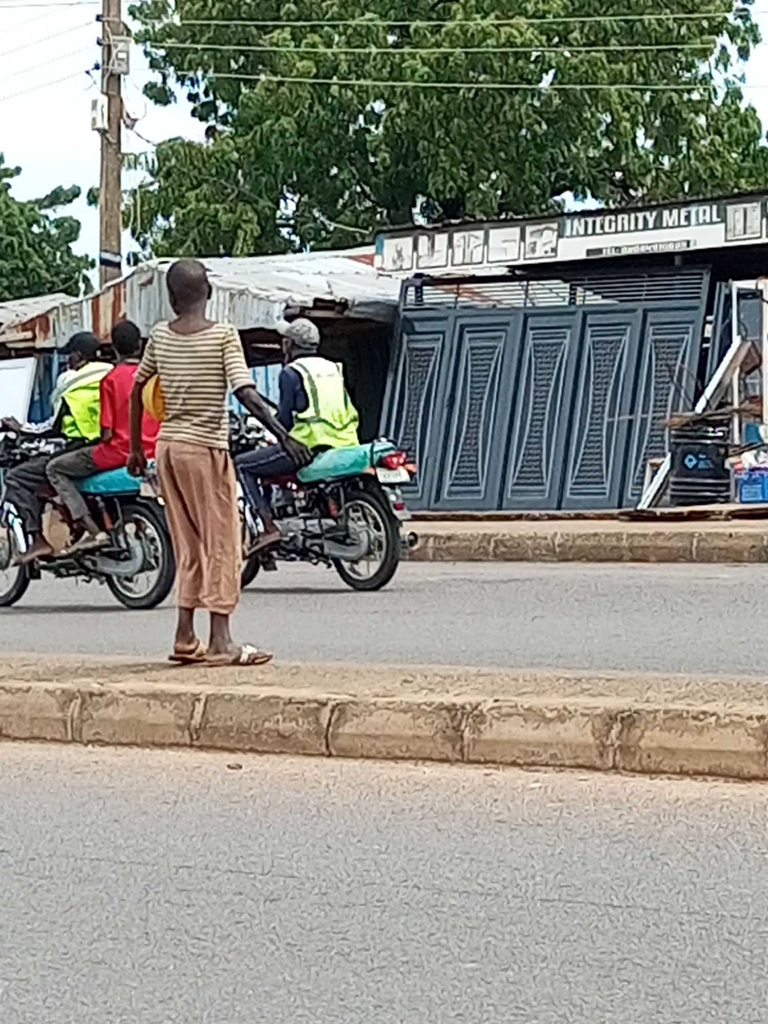 The image depicts a street scene in Garko, Nigeria, featuring a group of people on motorcycles. In the foreground, a person wearing a striped shirt and brown pants stands on the sidewalk. Behind them, two motorcycles are visible. The first motorcycle has two passengers wearing high-visibility vests, and the second motorcycle has one passenger also wearing a high-visibility vest. In the background, buildings with corrugated metal roofs are present, along with a sign that reads "INTEGRITY METAL". The environment appears to be an urban or semi-urban setting, with trees and overhead power lines contributing to the scenery. The road is paved.