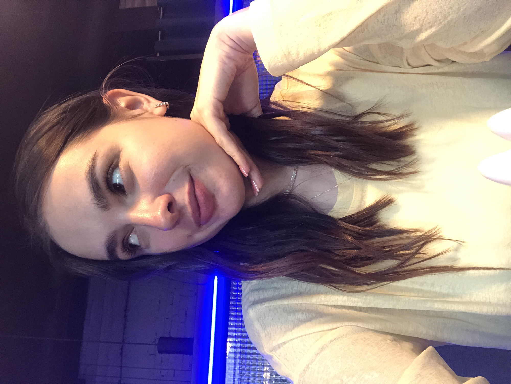The image features a young woman, likely in her late teens or early twenties, taking a selfie. She has long, dark brown hair and appears to be resting her head on her hand, with her fingers gently touching her forehead and temple. She is wearing a light-colored, long-sleeved top and a delicate necklace. A ring is visible on her right ear.
The background is dark and out of focus, but suggests an indoor setting with what appears to be a blue light source casting a glow on the left side of the image. The lighting is warm and soft, highlighting the woman's features. Her expression is relaxed and contemplative, with a slight smile.
The image seems to be taken at night or in a dimly lit environment, possibly at home or in a casual social setting. There is no visible text in the image. The overall mood is intimate and personal.