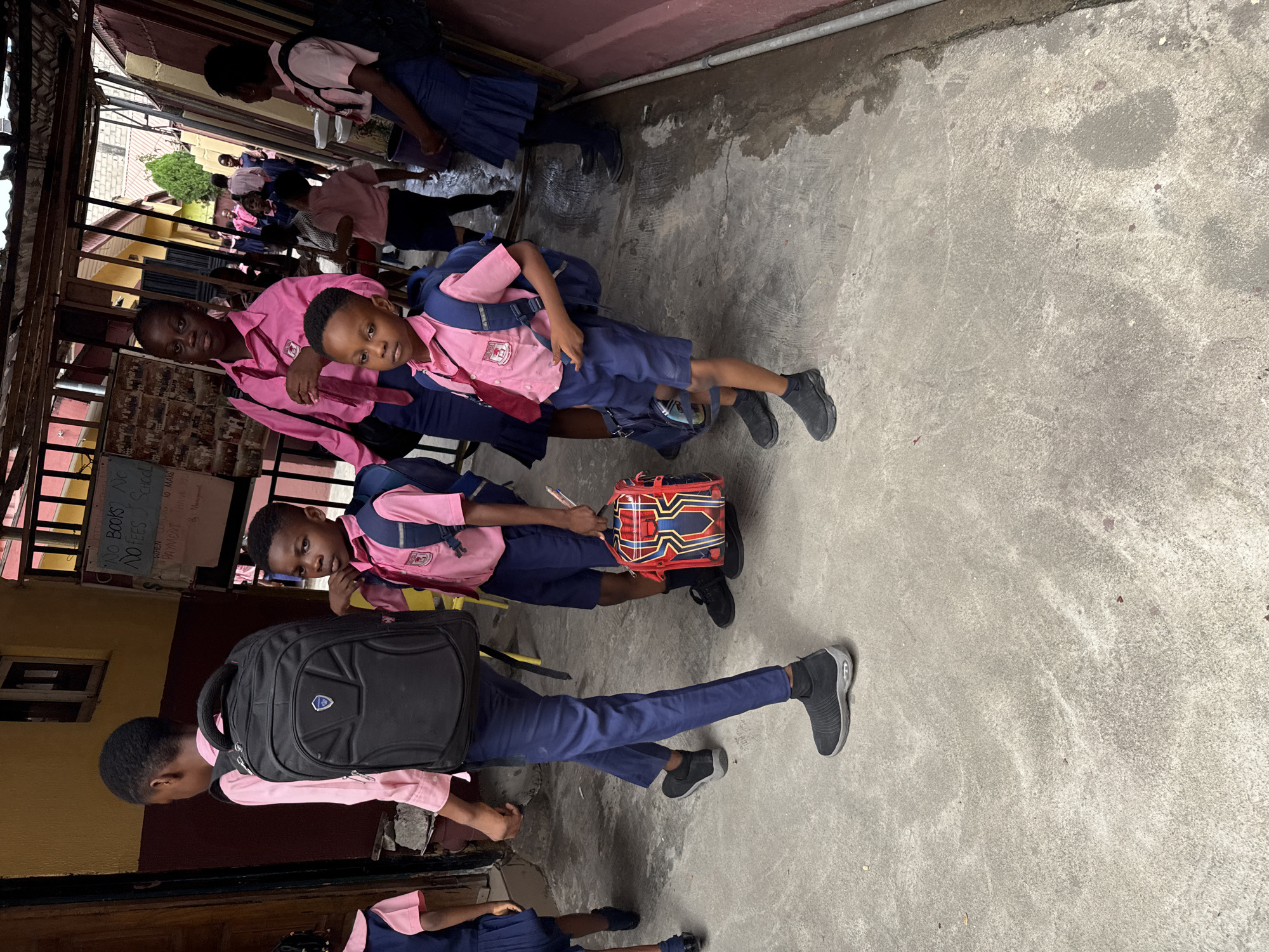 A group of school children in pink shirts and blue trousers or skirts are standing in front of a building with a sign reading "NO BOOKS NO FEES SCHOOL, NO FEES SCHOOL". The environment appears to be a concrete walkway or courtyard with some vegetation visible in the background. The location is Ado, Nigeria, based on external information. The children are wearing backpacks and appear to be waiting or gathering together.