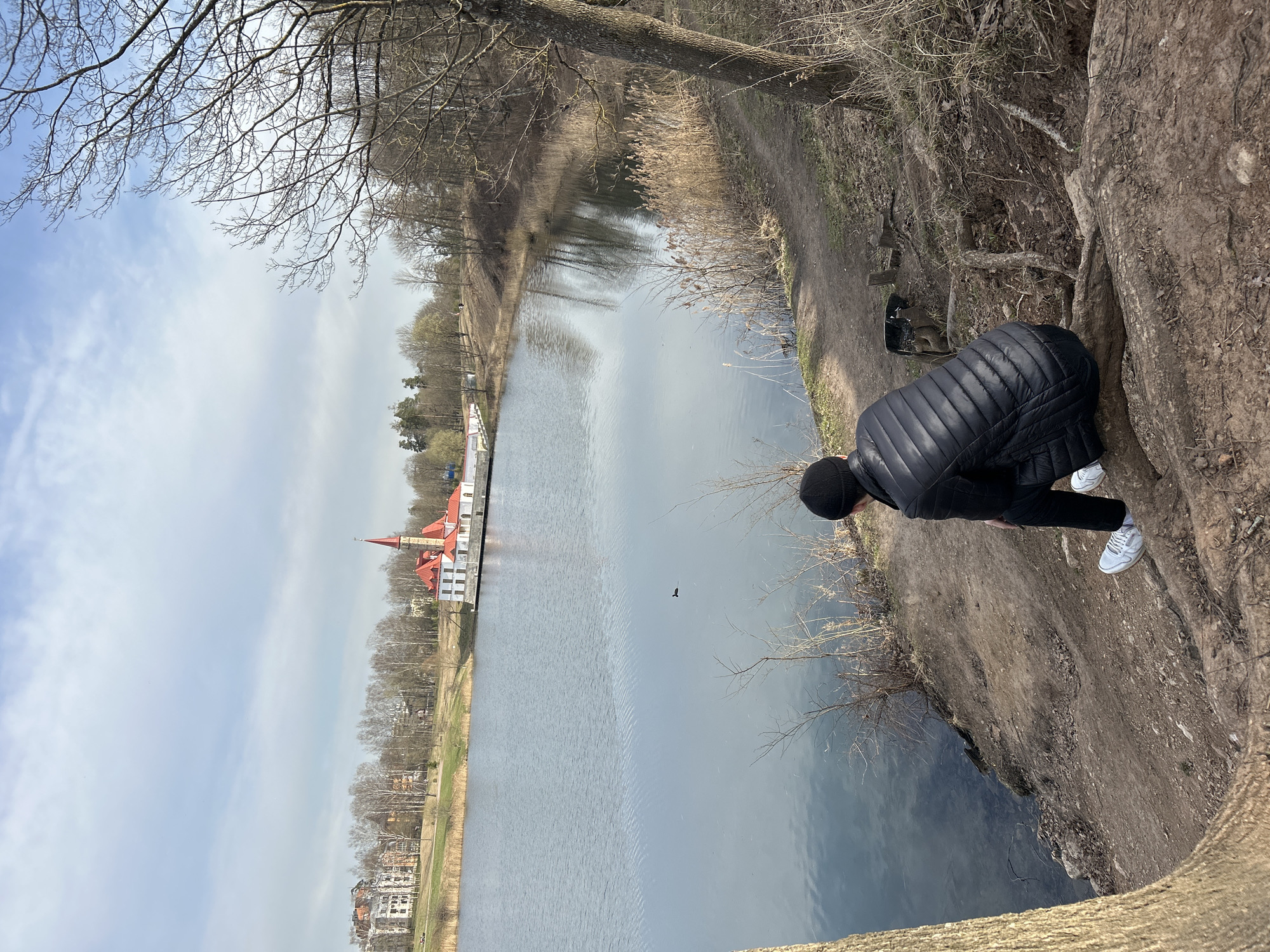 The image shows a person standing on the edge of a riverbank looking out over a body of water towards a building with a red roof. The person is wearing a black jacket, black pants, a black beanie, and white sneakers. The riverbank is made of dirt and has some vegetation growing on it. The building on the other side of the water appears to be a long structure with multiple sections, some with white walls and a red roof. The sky is overcast, and there are trees visible in the background. The location seems to be a park or natural area within a city.