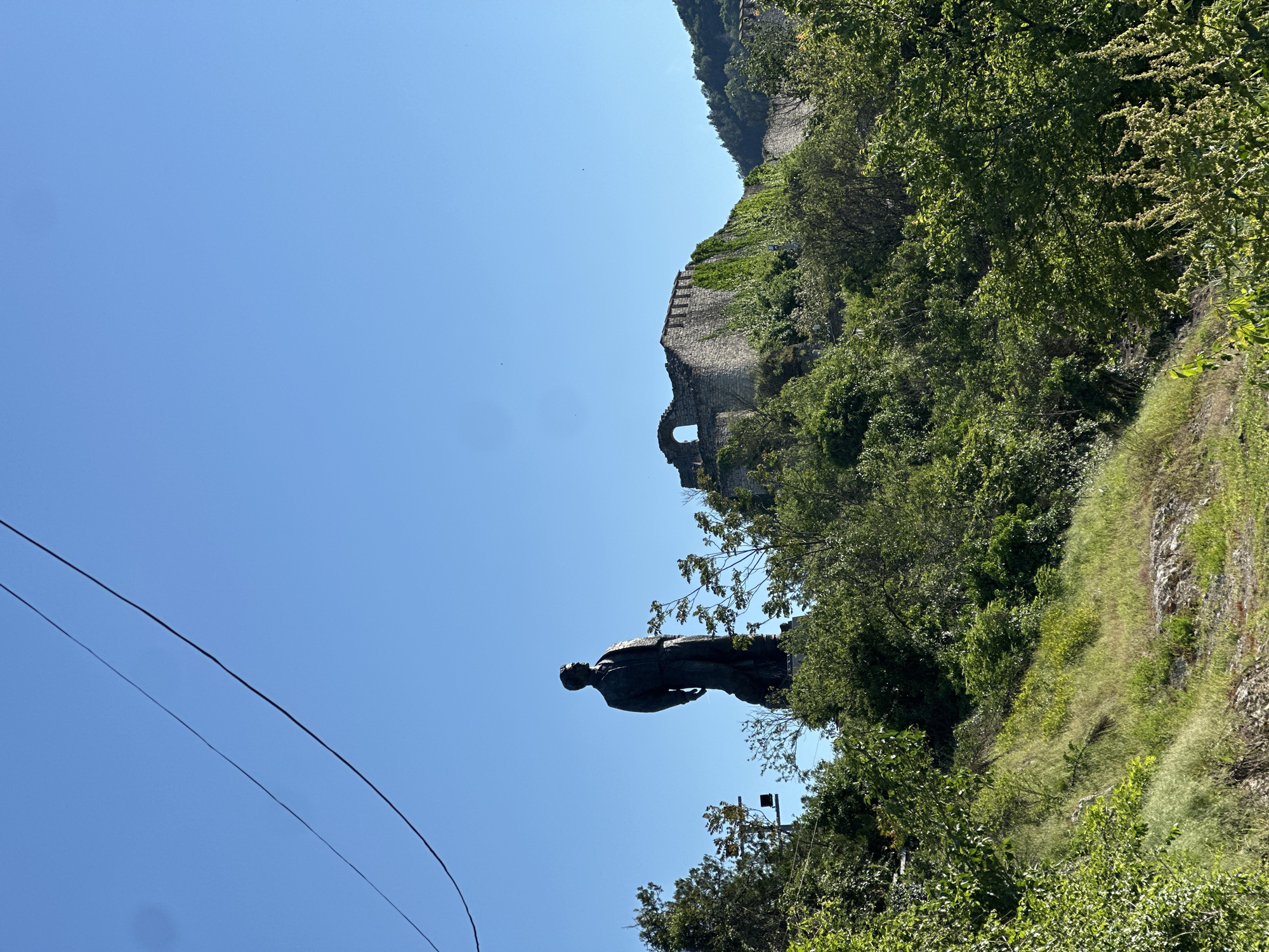 The image captures a statue positioned on a slope, with a medieval fortress visible in the background. Lush greenery surrounds the statue and the fortress. Above, a clear blue sky fills the frame. A power line crosses the bottom left of the scene. The location is Lovech, Bulgaria.