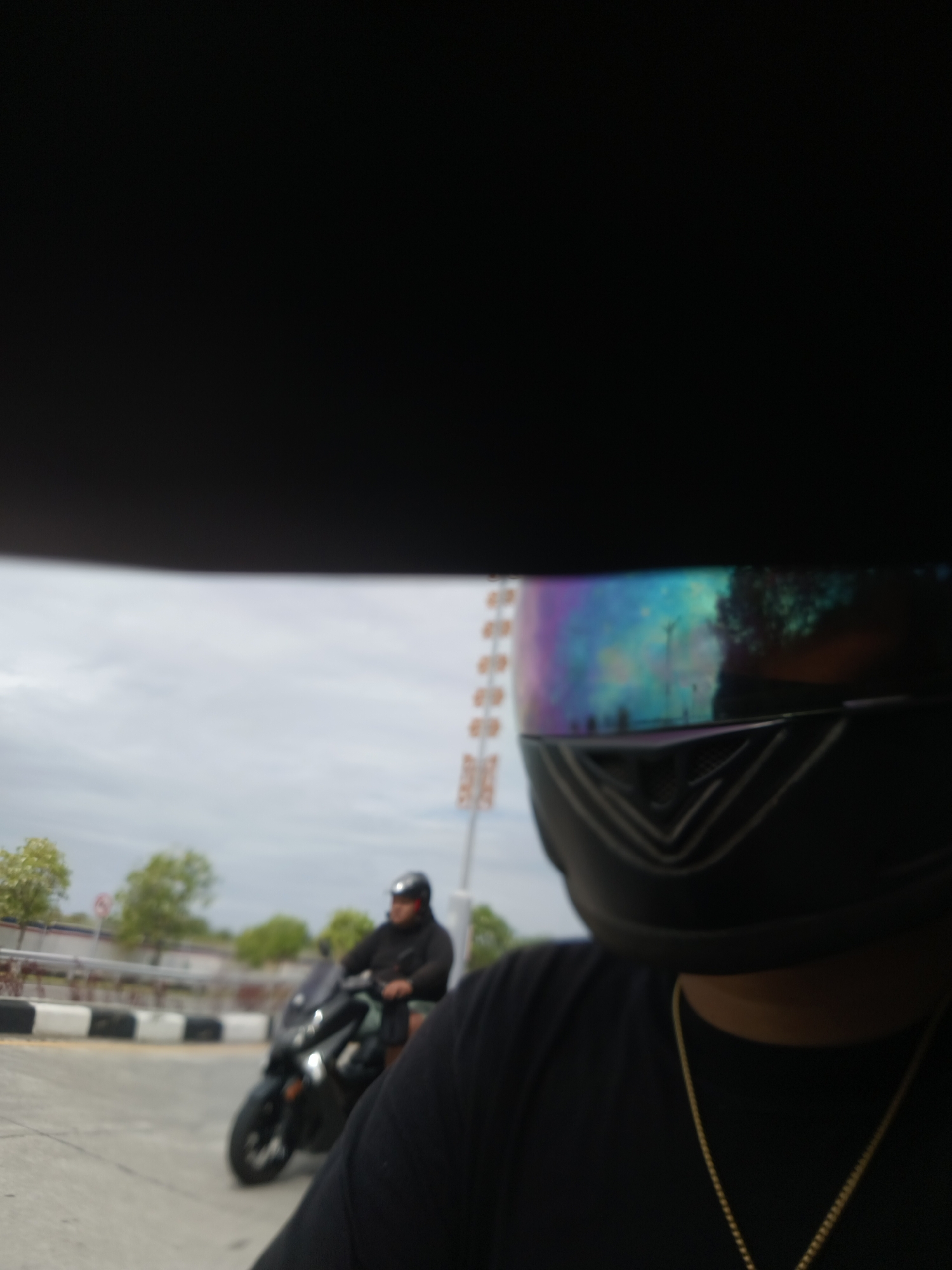 In Denpasar, Indonesia, a person wearing a black t-shirt and a motorcycle helmet with a reflective visor is seen from the chest up. The visor displays a colorful reflection of the sky and trees. In the background, another person is riding a black scooter. The scene appears to be outdoors, possibly near a road, with a cloudy sky and some greenery visible.