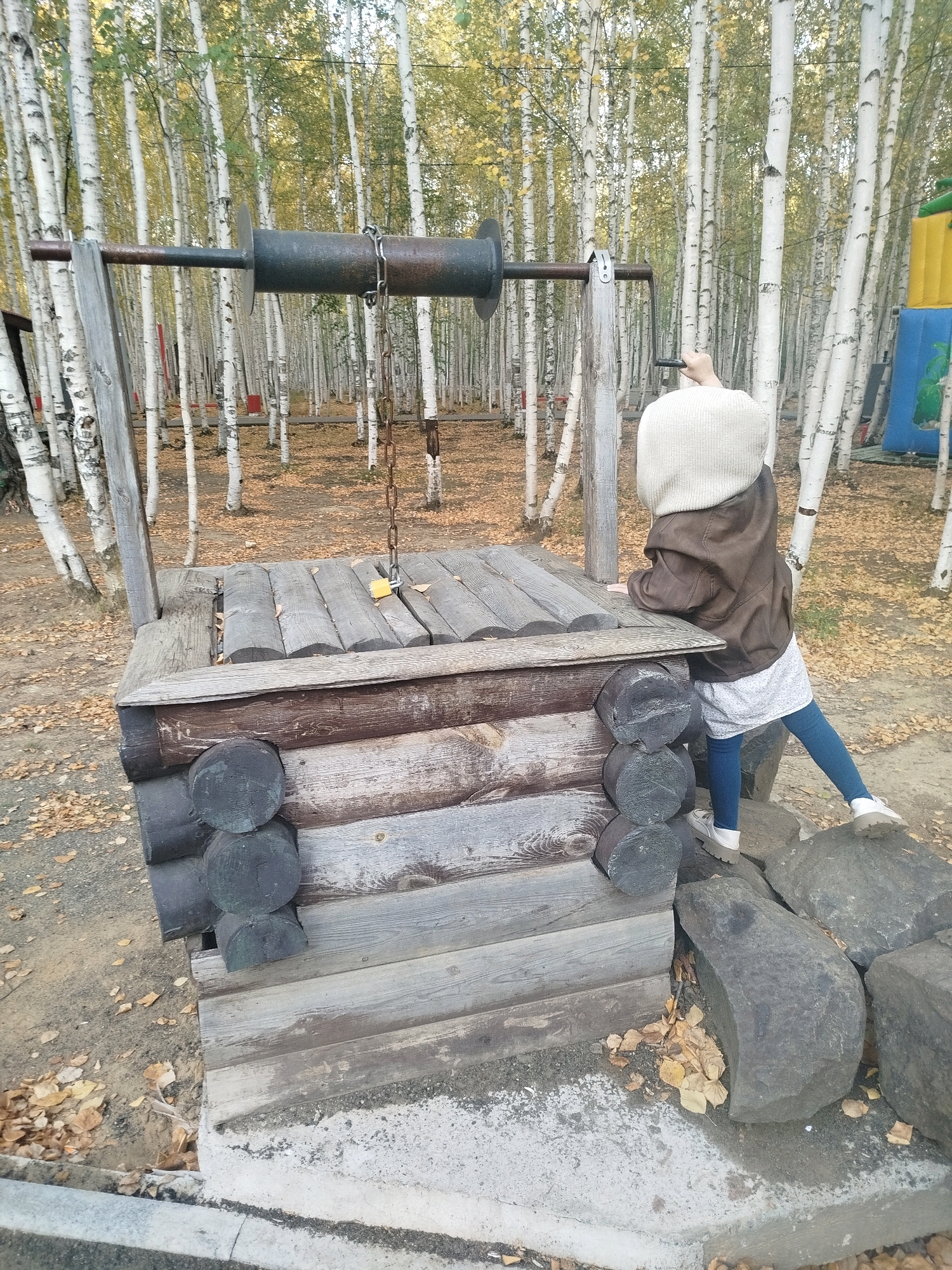 A child stands next to a rustic wooden well structure in an outdoor area, possibly a park or playground. The well features a wooden frame, a wooden lid, and a metal crank with a chain. The child, facing away from the camera, is wearing a beige knitted hat, a brown jacket, a white shirt, and blue pants with white shoes. They are reaching towards the well's mechanism. The background consists of a grove of birch trees with yellowing leaves, indicating an autumn setting. The ground is covered with fallen leaves and dirt. In the background, a colorful inflatable structure is partially visible, suggesting the presence of recreational facilities. The overall scene depicts a child engaged with an old-fashioned well in a natural, wooded environment. The exact location in Mirny, Russia, cannot be determined from the image alone.