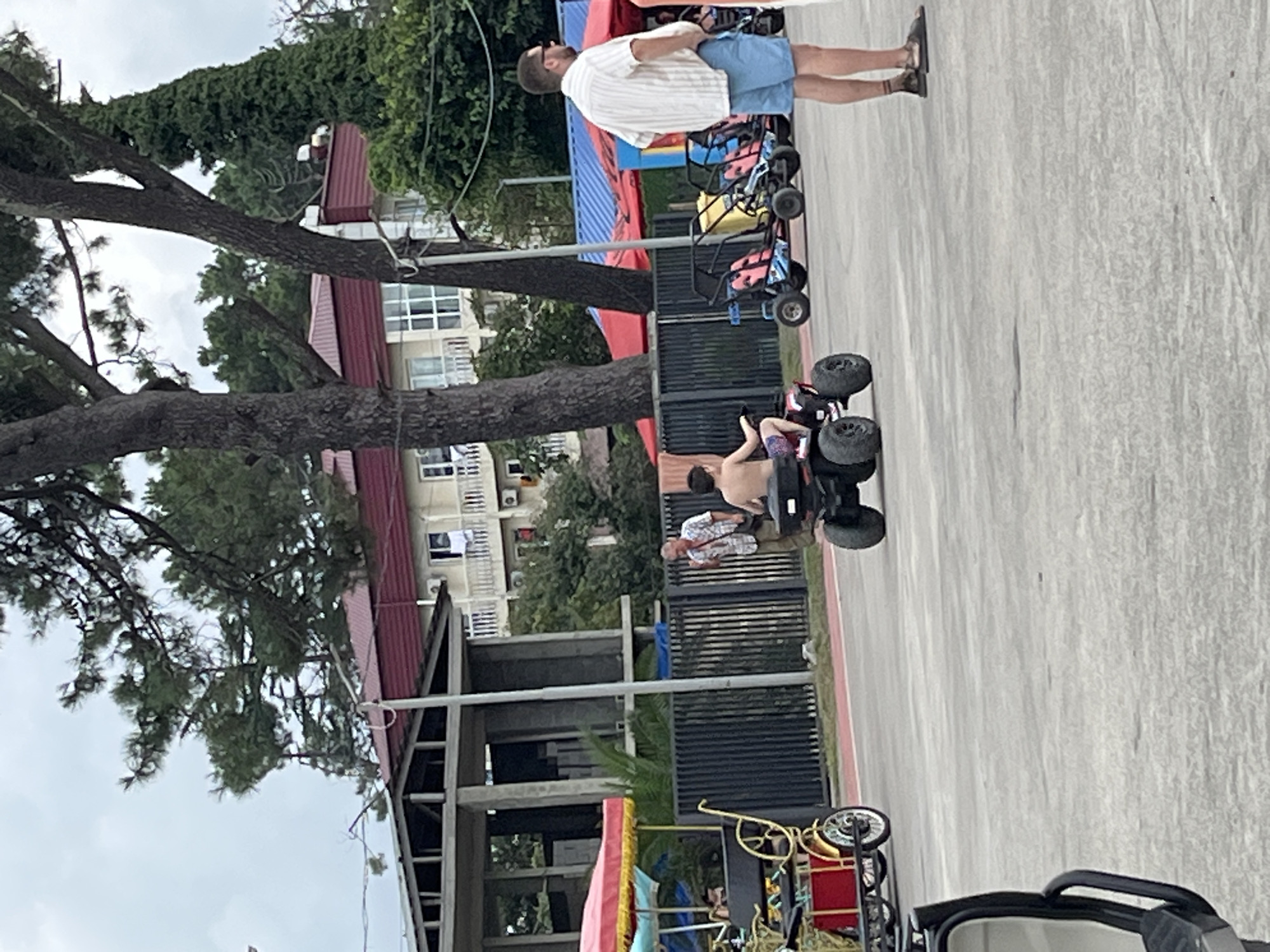 The image captures a scene in Ureki, Georgia, under an overcast sky. In the foreground, a young boy is operating a red and black quad bike. To his left, an adult male in a checkered shirt and khaki pants stands and gestures with his right hand. In the middle ground, a man wearing a striped white shirt and blue shorts stands with his hands on his hips, facing away from the camera. Behind him, a row of colorful multi-seater go-karts are parked. A yellow and red rickshaw-style pedal vehicle is partially visible on the far left. In the background, a multi-story building with a red roof is situated amongst trees. A partially constructed building with exposed concrete and beams is located to the left of the main building. A black metal fence separates the paved area from the greenery behind it.