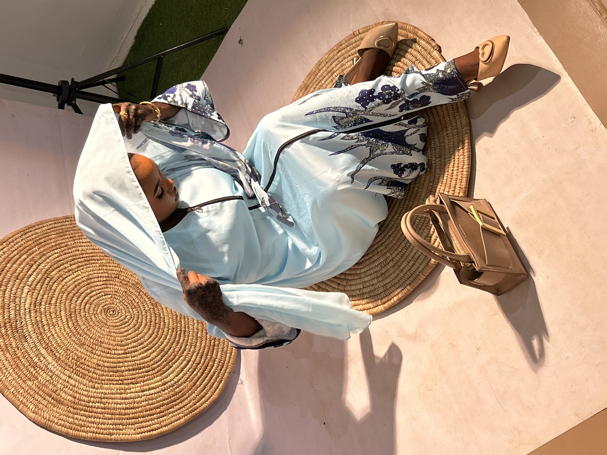 A light-skinned person is lying on a round, woven mat in what appears to be an indoor studio. The person is wearing a light blue outfit with dark blue patterned sleeves and pants, and beige shoes with a circular decoration. The person's head is covered with a light blue headscarf. A brown handbag is placed on the floor beside the mat. The floor and walls are a light beige color. Another similar round woven mat is positioned on the left side of the frame. There is an artificial grass patch and a photography light stand are visible in the background. Given the studio setting and the subject's attire, this might be a staged photograph for fashion or cultural representation. There is no definitive way to determine if the location is in Jos, Nigeria from just this single picture.