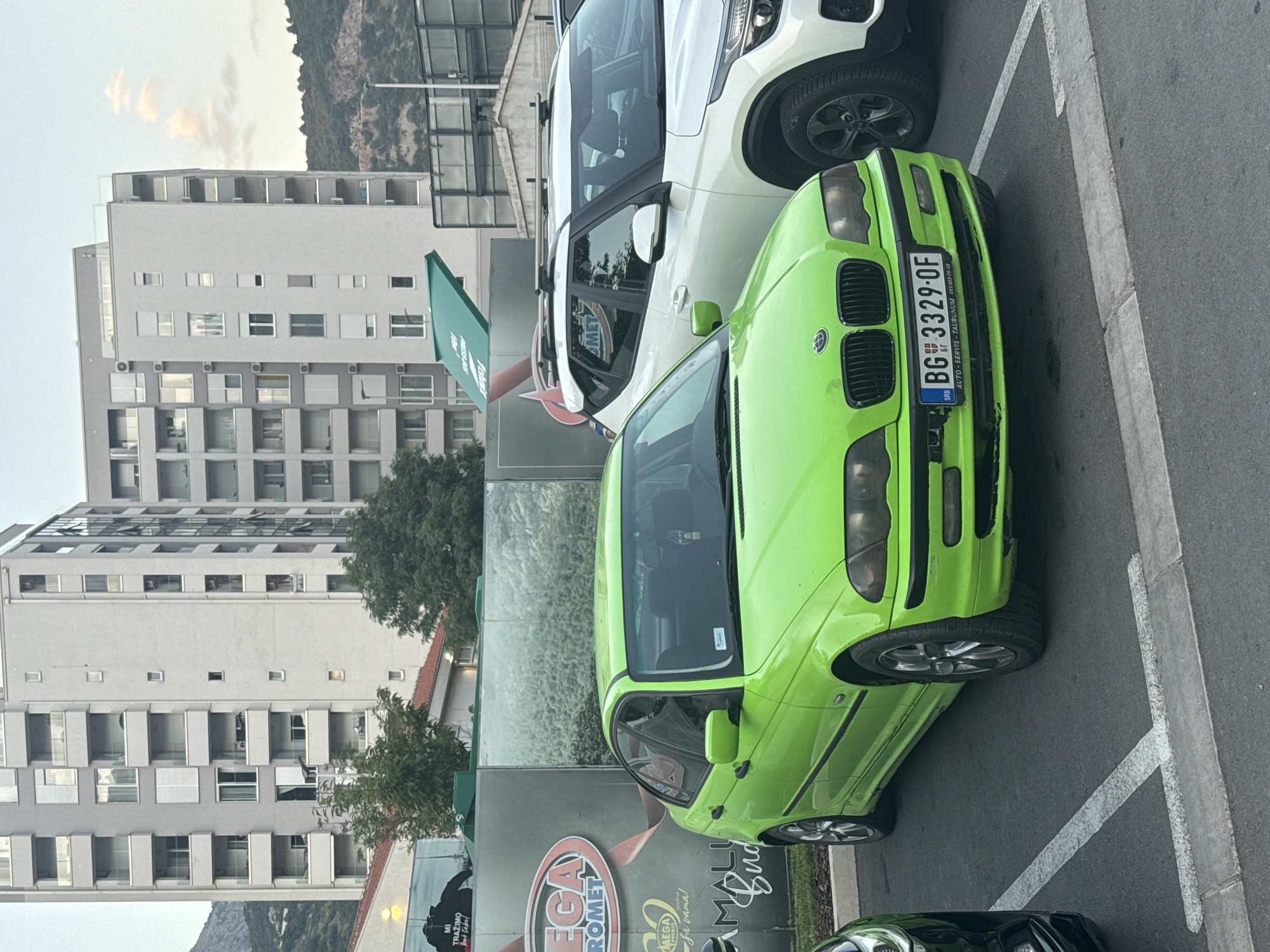 In Budva, Montenegro, a lime green BMW sedan is parked in a lot with modern apartment buildings in the background. The BMW's license plate is BG 3329-OF, and it has a black trim around the headlights and grille. Next to the BMW is a white SUV with a roof rack. In the background, a large advertisement featuring the word "MEGA ROMET" and "Budva" is visible. The sky is overcast, and a few clouds are present. The parking lot appears to be paved and lined with parking spaces. The overall scene depicts a typical urban environment in Budva.
