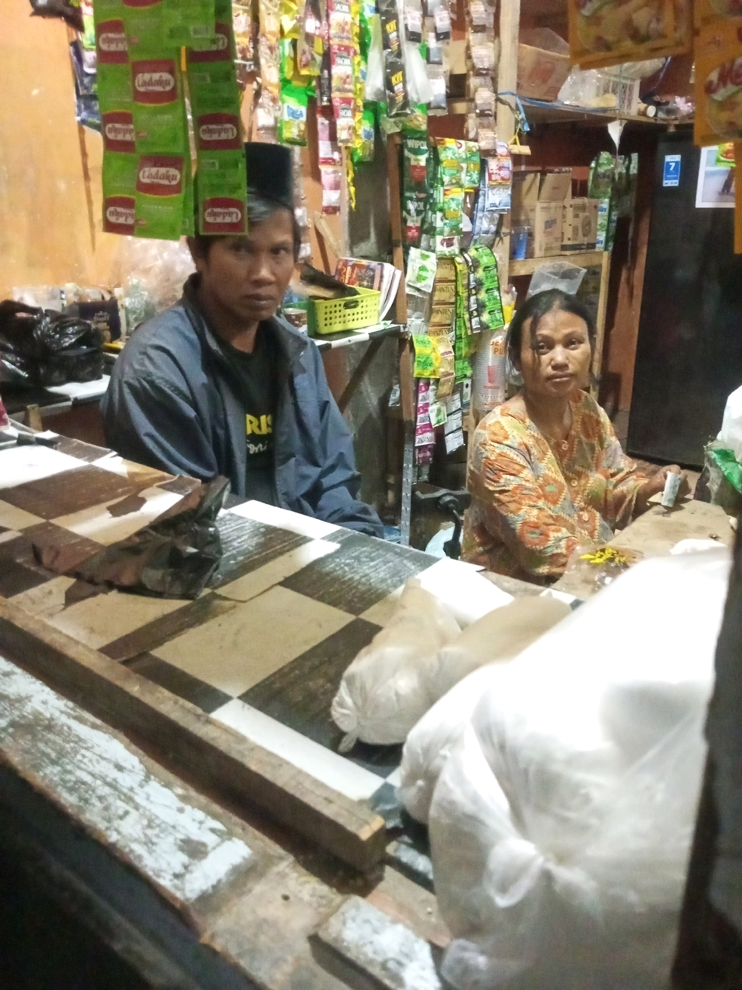 The image depicts a small, cluttered shop in Andongsili, Indonesia. A man wearing a black peci hat and a blue jacket is seated behind a checkered counter. To his right, a woman in a colorful patterned blouse is also behind the counter, holding a small white packet. The shop is filled with shelves stocked with various food items and other goods, predominantly packaged snacks and seasonings, hanging from the ceiling and walls. A refrigerator is visible in the background. In the foreground, large white plastic bags are stacked on the counter, partially obscuring the checkered surface. The lighting is dim, suggesting an indoor setting, possibly during the evening.