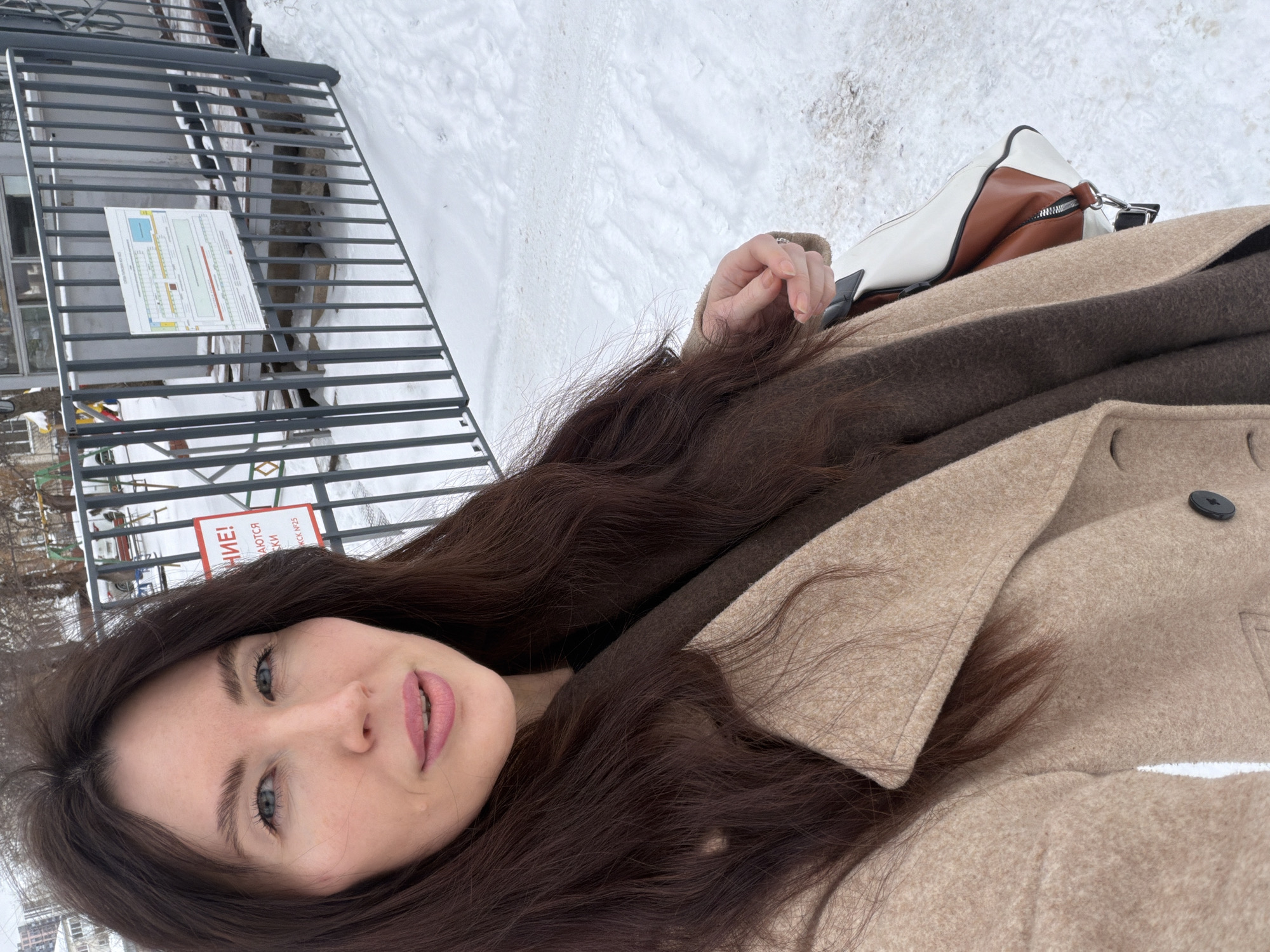 A woman takes a selfie outdoors in Perm, Russia, during winter. She is wearing a beige coat with a dark brown scarf, and has long, dark brown hair. A two-tone bag is visible next to her. A metal fence is behind her, with a sign attached to it. Snow covers the ground. The selfie appears to be taken in the city based on the background.