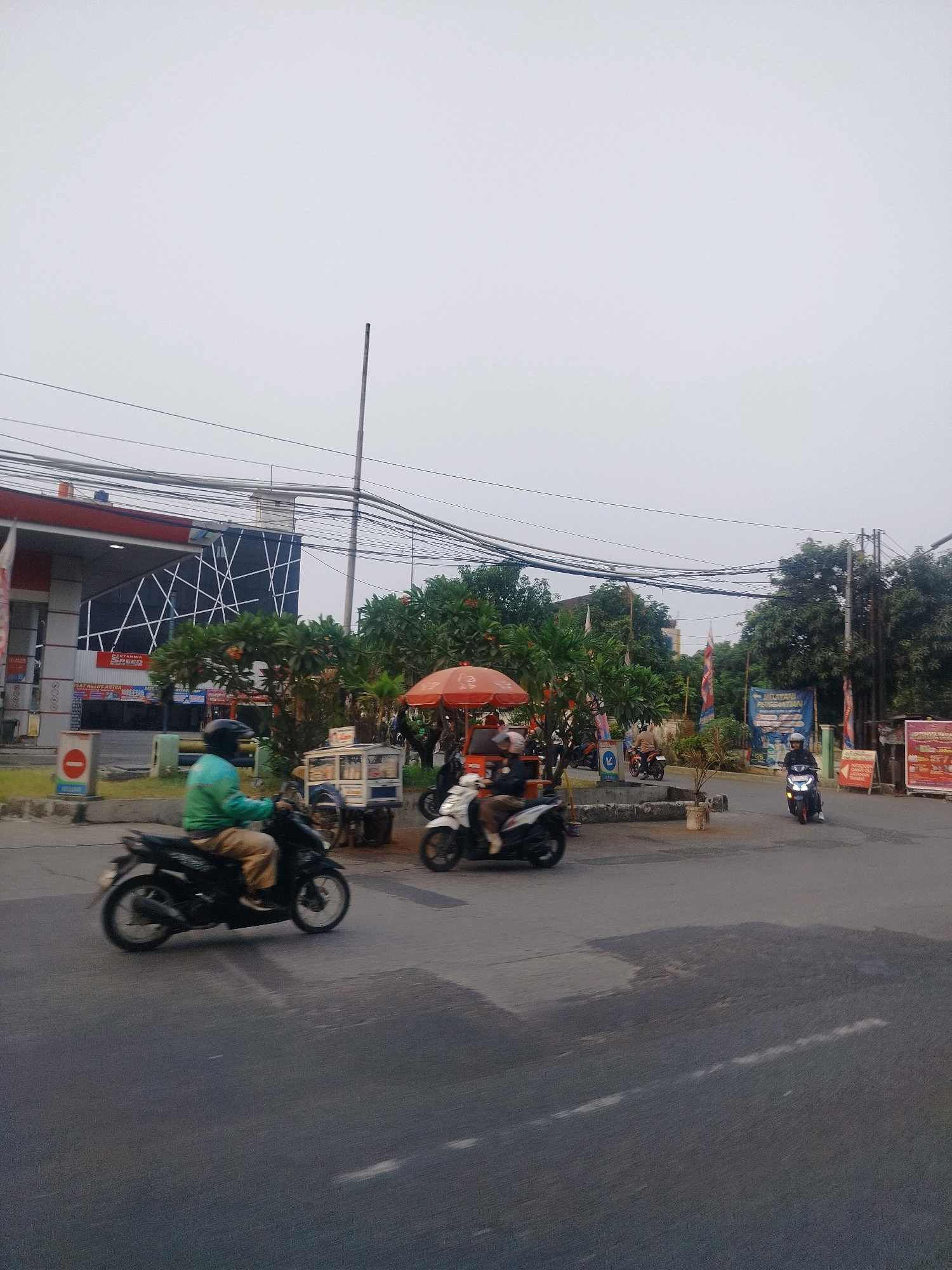 The image captures a street scene in East Jakarta, Indonesia, on an overcast day. A motorcycle, ridden by a person wearing a green jacket and beige pants, is in the foreground, moving from left to right. In the middle ground, several other motorcycles are visible. One is carrying two passengers, and another is ridden by a single person. A food cart with an orange umbrella is parked on the side of the road. Behind the food cart, a building with a modern facade, featuring a geometric pattern of white lines on a black background, appears to be a Pertamina gas station. Trees and utility poles with numerous wires are also present in the background, indicating an urban environment. Several signs are visible, including a red and white prohibition sign near the gas station. The road surface is asphalt, with some patches of wetness or discoloration. The overall impression is a busy street intersection in a residential or commercial area.