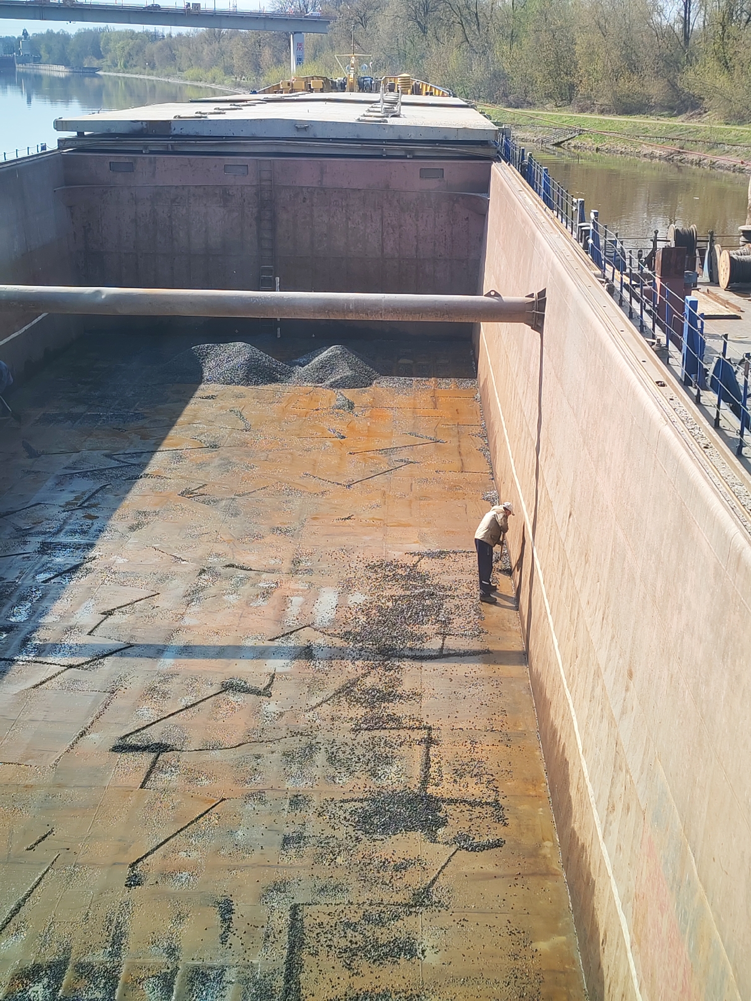An elevated, high-angle shot shows the interior of a large, rusted cargo barge containing loose gravel and an individual using a tool against the far wall of the barge. The gravel is piled near the center. A long, gray, horizontal pipe runs across the width of the barge at the top.
The barge is located in a canal or wide river; the background shows a riverbank with trees and a bridge. The location is Yakhroma, Russia, indicated by the bridge in the background.
The individual, dressed in dark trousers and a light-colored jacket, is positioned to the right in the image, interacting with the wall. Blue railings line the edge of the barge on the right side. The barge floor appears to be marked with square or rectangular segments. Shadows cast across the scene suggest a sunny day.