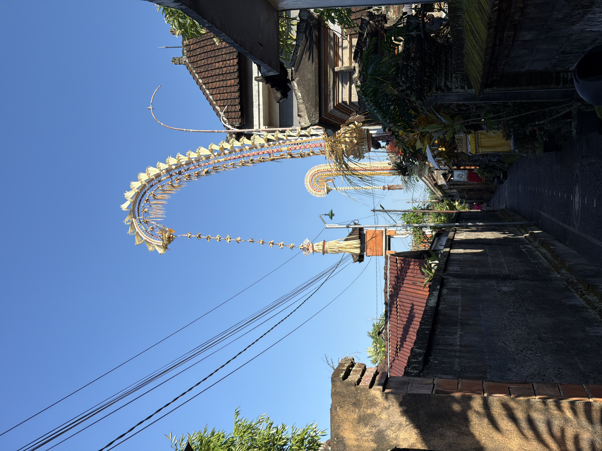 A narrow alleyway in the city of Ubud, Indonesia, is bordered by walls and buildings. Ornate, festive decorations, likely Penjor, are erected on either side of the alley, their curved forms reaching towards the clear blue sky. One Penjor, a tall, elaborately decorated bamboo pole, towers above the rooftops, adorned with golden accents and hanging ornaments. Another, a more curvilinear structure, also displays vibrant colors and traditional embellishments. Utility wires crisscross overhead, casting shadows on the aged walls. The architecture features terracotta tiled roofs and stone or concrete construction. Lush greenery is visible in some areas, adding to the tropical atmosphere. The alleyway itself is paved and appears to be a thoroughfare within a residential or cultural area. The scene is captured under bright daylight, suggesting a warm, sunny day.