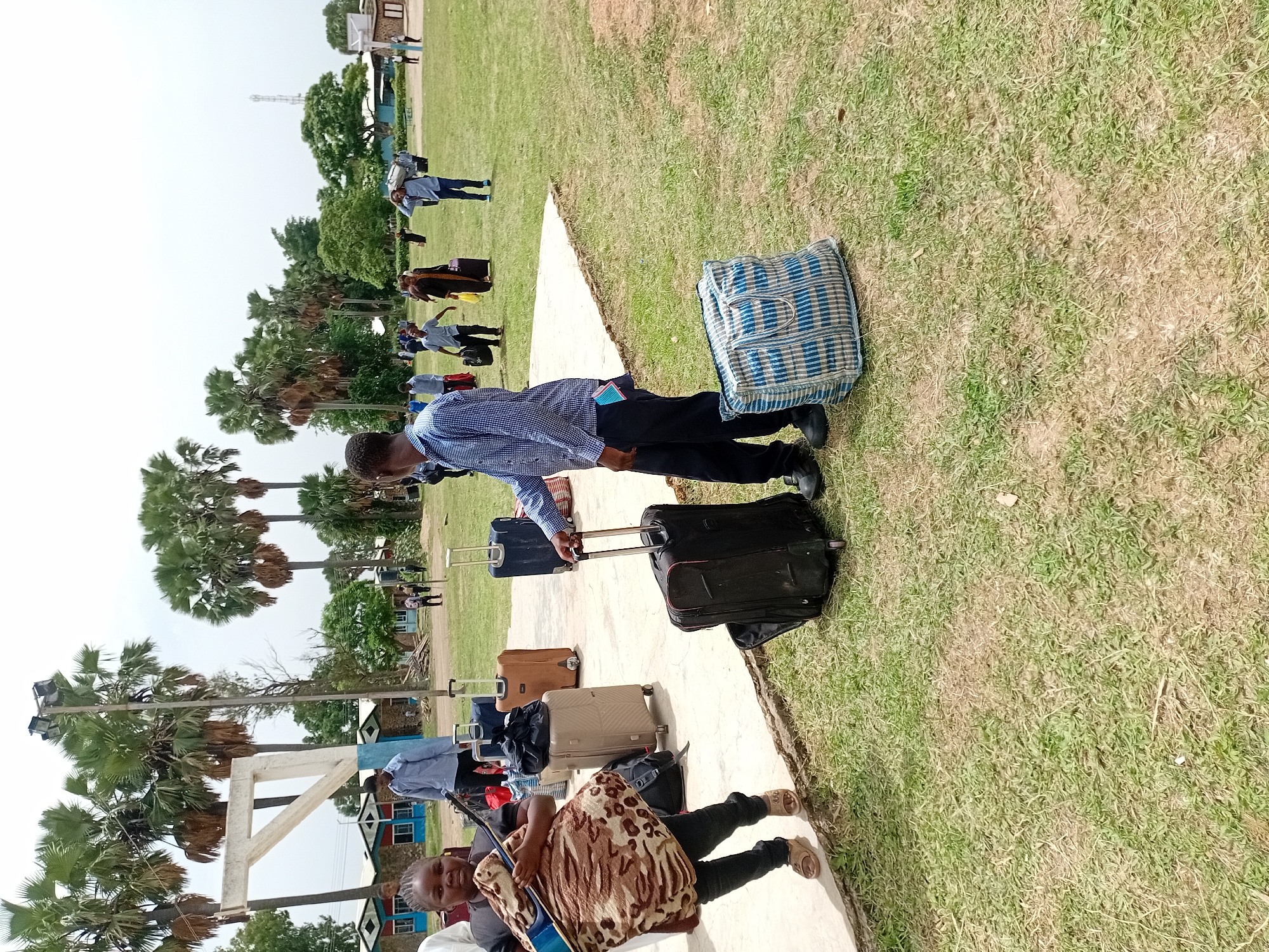 The image depicts a group of people standing and sitting alongside a concrete walkway, likely at an outdoor location. Many are carrying luggage, including suitcases and bags. A man in a blue patterned shirt is bent over, appearing to adjust a suitcase. A woman sits on the walkway holding a guitar. Trees are visible in the background. While it cannot be confirmed to be Jos, Nigeria with the provided visual information, the scene could be located anywhere.