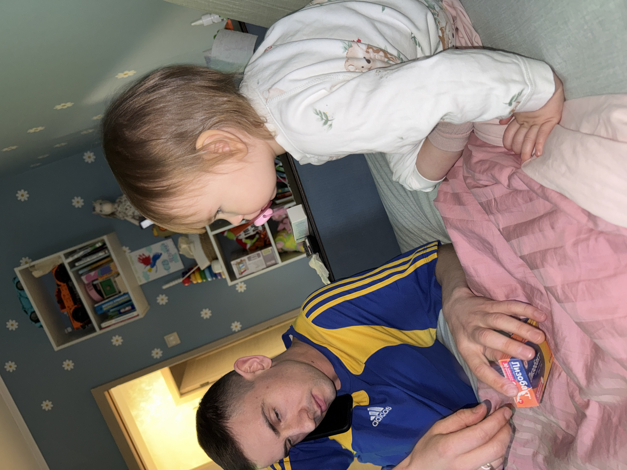 The image portrays a tender indoor scene, likely in a bedroom in Moscow, Russia. An adult male is seen resting or sleeping, lying down with his eyes closed. He is wearing a blue and yellow tracksuit with the "adidas" logo visible on the sleeve. A black cell phone is positioned near his head. In his hands, he holds a small orange and white box of medication, clearly labeled in Cyrillic "Лизобакт" (Lysobact) and the number "30".
Leaning over him from a higher surface, possibly a bed or crib, is a young child with light brown hair, wearing white pajamas adorned with animal and leaf patterns. The child has a pink pacifier in their mouth and appears to be observing the man. The bedding visible under and around the man is a light pink color.
The background wall is a soft blue-green, decorated with numerous small white daisy decals, suggesting a child's room. To the left, a white shelving unit is visible, holding various toys and books, including a blue car and an orange truck. The overall atmosphere is quiet and domestic, capturing a peaceful moment between a child and an adult.