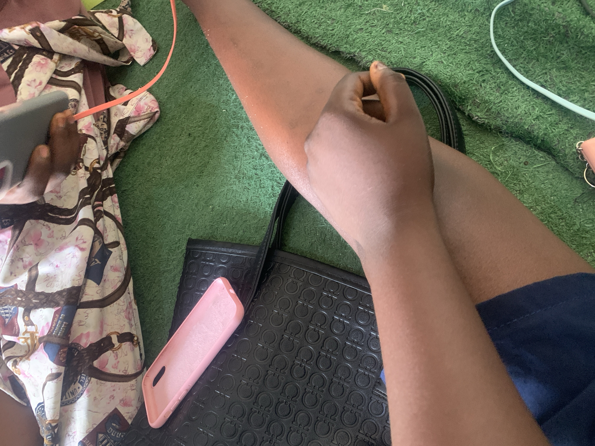 The image depicts a close-up view of two arms on a green, artificial turf surface, possibly a mat or carpet. The arm on the right is extended, with its skin showing a warm, slightly reddish tone, and appears to be resting. The arm on the left is bent, with a hand clasped over the forearm of the extended arm. Both individuals' skin tones appear to be dark.
On the left side of the image, a black textured clutch or bag is visible, with a pink phone case resting on top of it. The bag has a repeating pattern of embossed logos. In the upper portion of the image, a piece of fabric with a busy pattern of buckles, straps, and floral motifs is draped. A grey mobile phone is partially visible, connected to an orange charging cable that snakes out of frame. A light blue cable is also visible in the lower right corner.
The overall scene is one of casual resting or interaction, with personal items scattered around. The location is identified as Jauro Yinu, Nigeria. The artificial turf suggests an indoor or possibly an outdoor setting designed for recreational use, common in urban areas. There are no visible buildings or specific city landmarks that would provide further detail about the precise location within Jauro Yinu. The lighting appears to be natural, suggesting daylight.