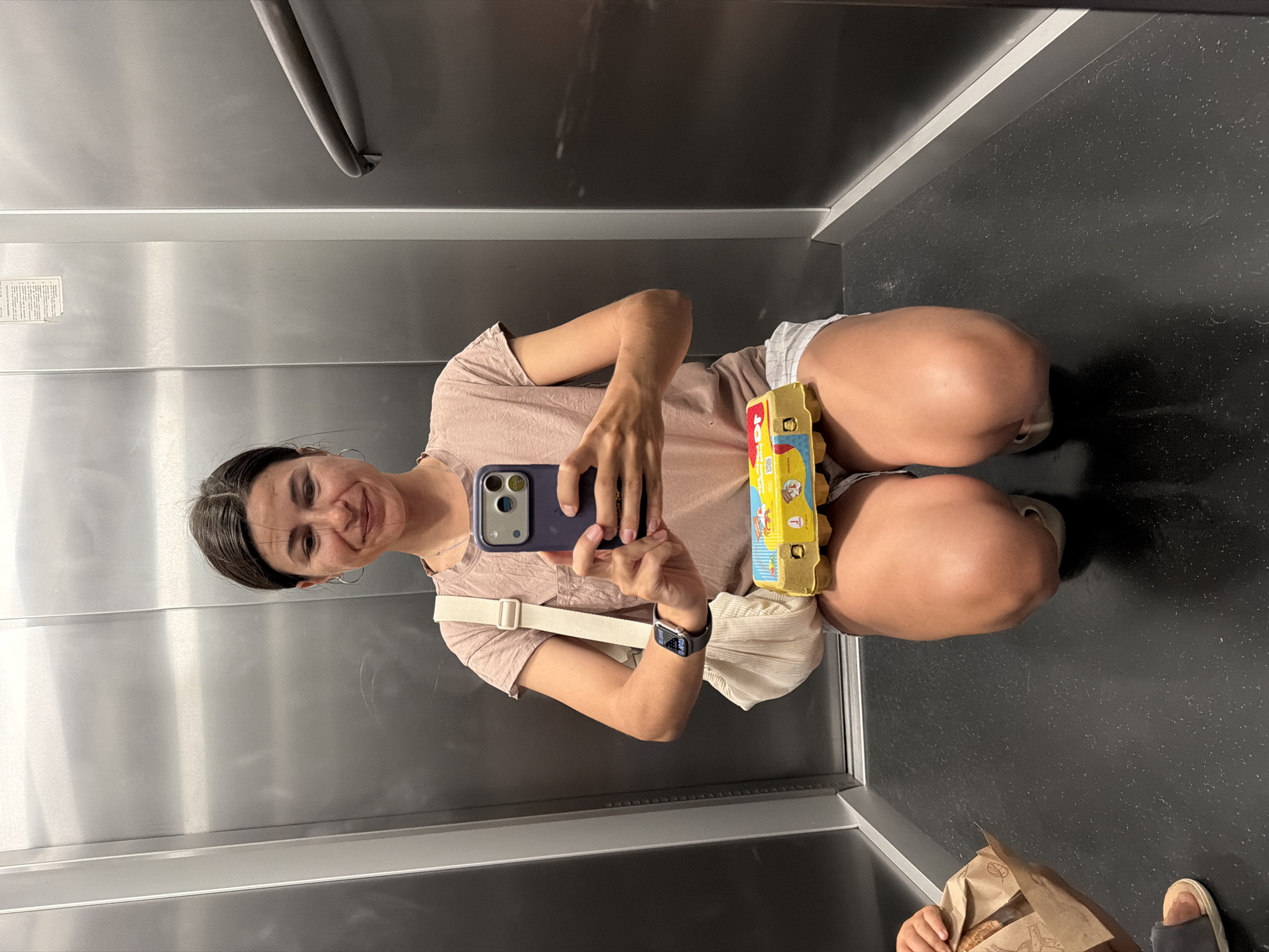A woman is taking a mirror selfie in an elevator, likely in Budva, Montenegro, based on the presence of a product from the "Franc" bakery chain. The woman is holding a carton of eggs in one hand and her smartphone in the other. She is wearing a casual outfit consisting of a light pink t-shirt and white shorts. She is also wearing a watch on her left wrist and a crossbody bag. Another person's hand holding a brown paper bag from the Franc bakery is visible in the lower right corner of the image. The elevator has brushed metal walls and a dark gray floor. A horizontal handrail is visible on the upper left.