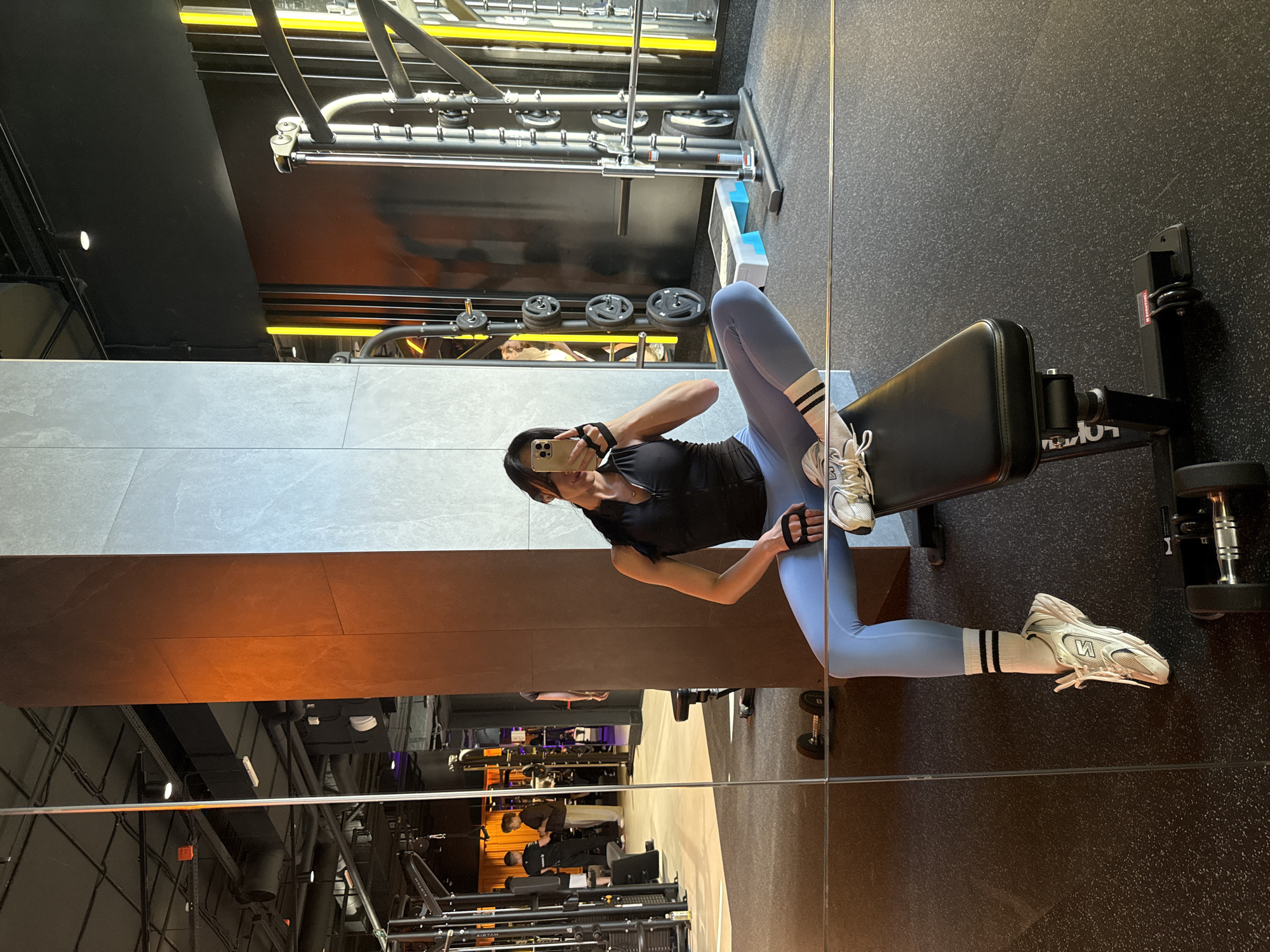 A woman in blue leggings and a black shirt is sitting on a weight bench in a gym, taking a selfie in a mirror. The gym equipment, including weights and machines, is visible in the background. The gym appears to be in Krasnoyarsk, Russia.
