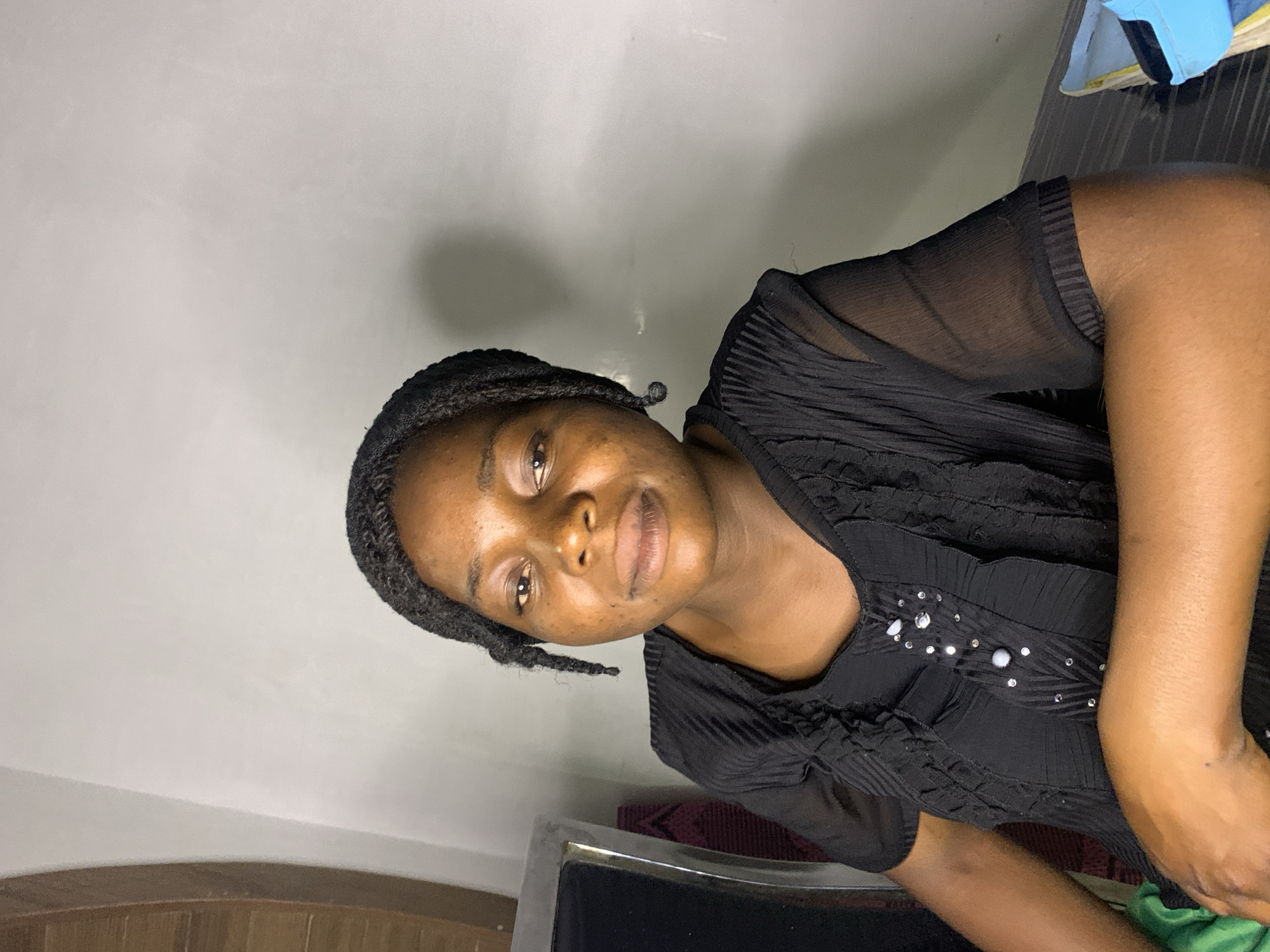 A medium shot captures a woman from the chest up, seated indoors with a pale gray wall as the background. She is facing slightly to the right, with her head tilted and her gaze directed forward. Her dark hair is braided into two strands that hang down. She is wearing a black V-neck shirt with a ruffled texture and decorative embellishments, including clear rhinestones and white buttons, down the front. Her left arm rests on her lap, and her right arm is bent at the elbow, with her hand resting on her chest. The lighting creates a soft shadow on the wall behind her, suggesting the light source is to her left. The setting appears to be an indoor space, possibly a living area or office, but specific details about its location within Jos, Nigeria, are not discernible from the image.