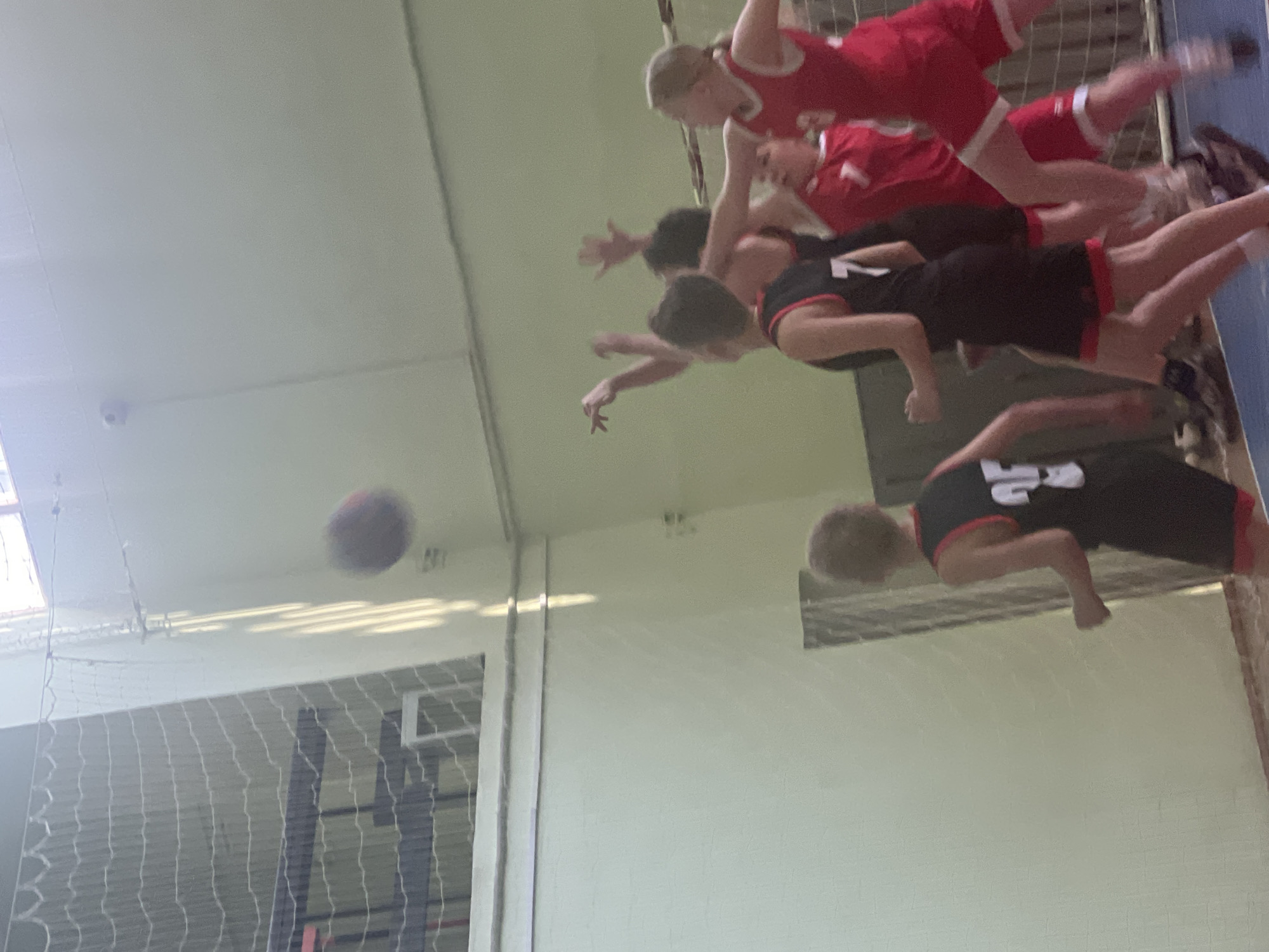 The image depicts a group of people playing basketball indoors in a gymnasium with white walls and a high ceiling in Chaikovsky, Russia. One person is in mid-air, presumably attempting a shot or rebound, wearing a red uniform with the number "7" on it. Other players, some in red uniforms, and some in black and red uniforms, are positioned below, seemingly engaged in the game. A basketball is visible in the air near the basketball hoop.