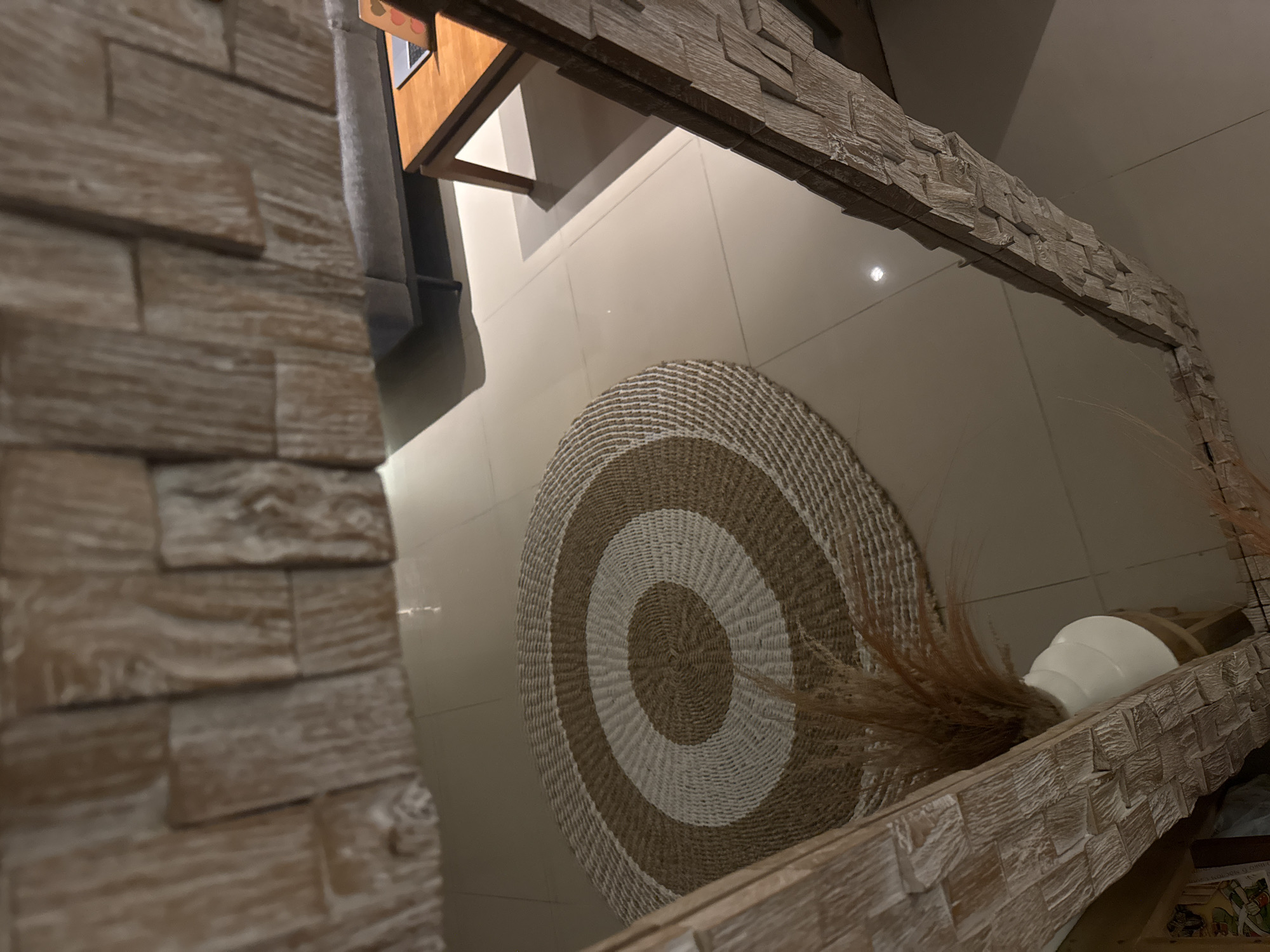 The image shows a room in Kuta Selatan, Indonesia. The foreground features a textured, light brown brick wall. In the center, a circular rug with alternating brown and white concentric stripes lies on the floor. A white vase is partially visible on the right edge, with dried foliage extending from it. Reflected in a mirror in the top right, is a section of the room's interior. A wooden table with a gray object on it is reflected in the upper left. The floor appears to be tiled with light beige tiles. The scene is well-lit.
