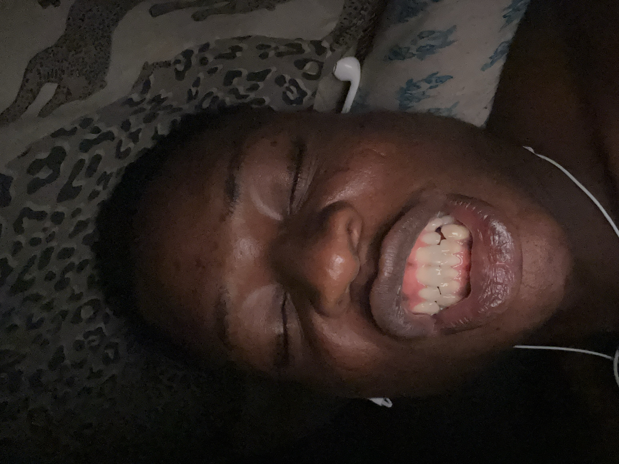 A close-up image depicts a person lying down, face upward, with eyes closed and mouth slightly open revealing teeth. The person has dark skin and a visible earbud in one ear. A white wire, likely from the earbud, is also visible. The person is lying on a surface with a patterned fabric. The location and context are not specified.