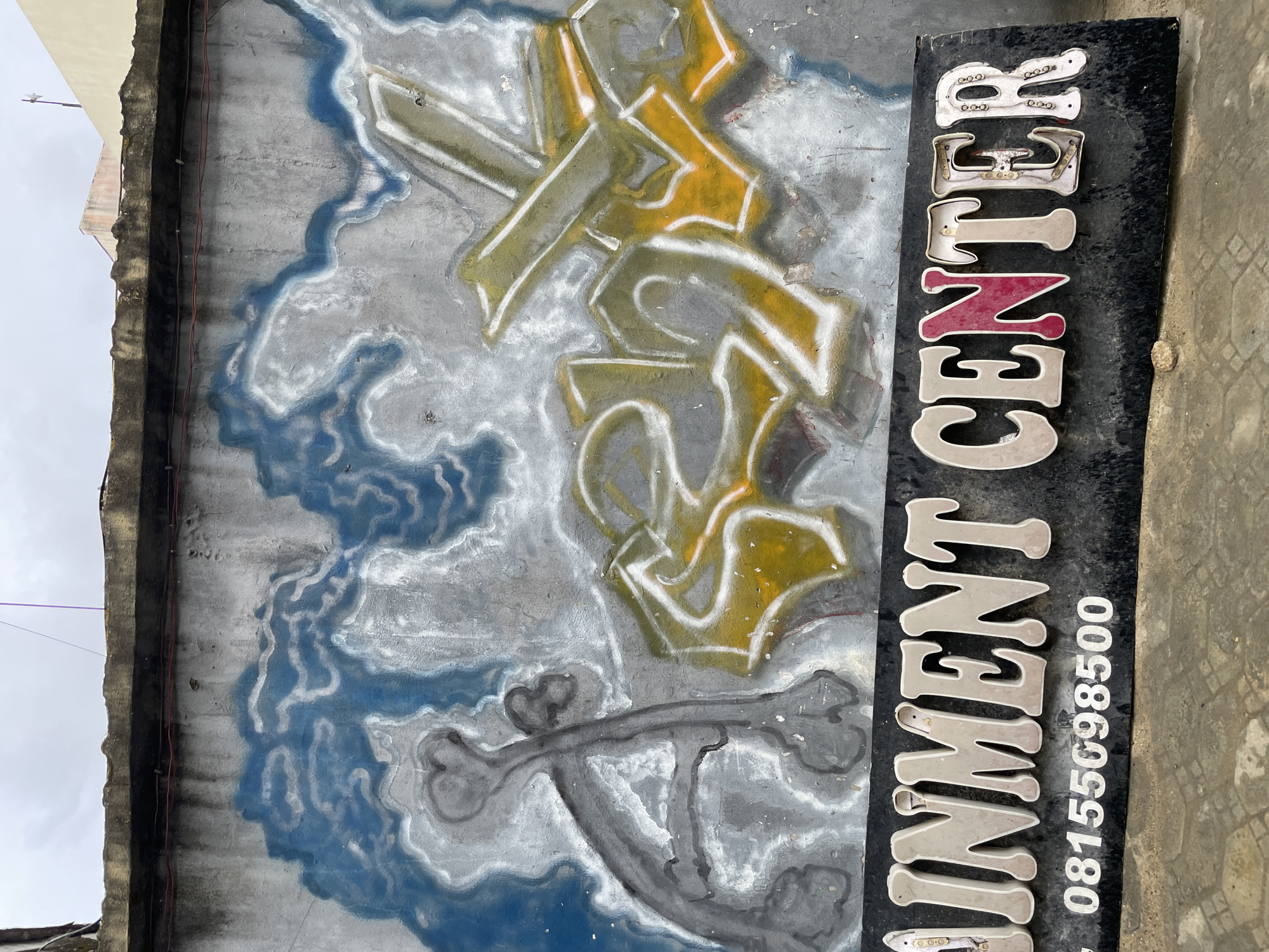 This is an exterior, eye-level, medium shot of a graffiti-covered wall in Ado, Nigeria. The graffiti art appears abstract, with blue and white swirls on the upper portion of the wall, and what looks like stylized text in green, yellow, and white in the center. Below the graffiti, a black sign with white and pink lettering reads "AINMENT CENTER" followed by a phone number "08155698500". The wall is rough and textured, showing signs of age and wear. The ground in front of the wall is paved with concrete or stone. The sky is overcast.