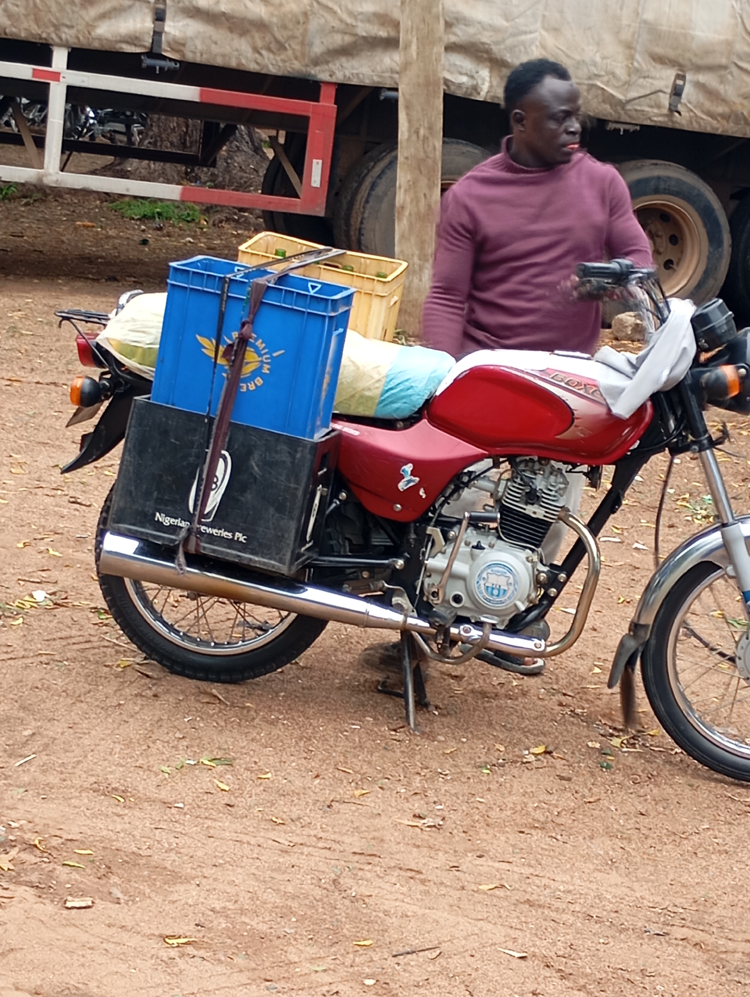 A man stands next to a red motorcycle, looking to the right. He wears a burgundy long-sleeved shirt. The motorcycle is parked on a dirt surface with some scattered leaves. On the back of the motorcycle are two crates, one blue and one black, both secured with straps. The blue crate has yellow lettering and a yellow emblem with wings. The black crate has white lettering that reads "Nigerian Breweries Plc." Behind the motorcycle and man is a large truck with a tan tarp covering its cargo. The environment appears to be outdoors, possibly a street or marketplace.